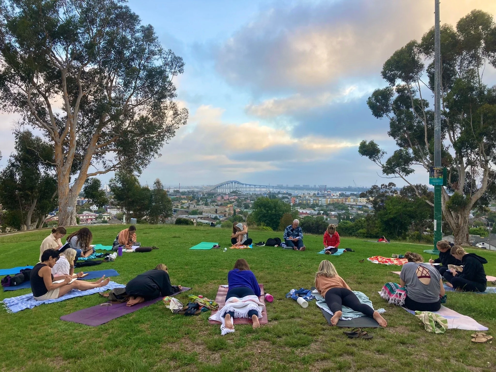 Outdoor yoga class on a grassy hill overlooking a city, bay, and large arched bridge at sunset