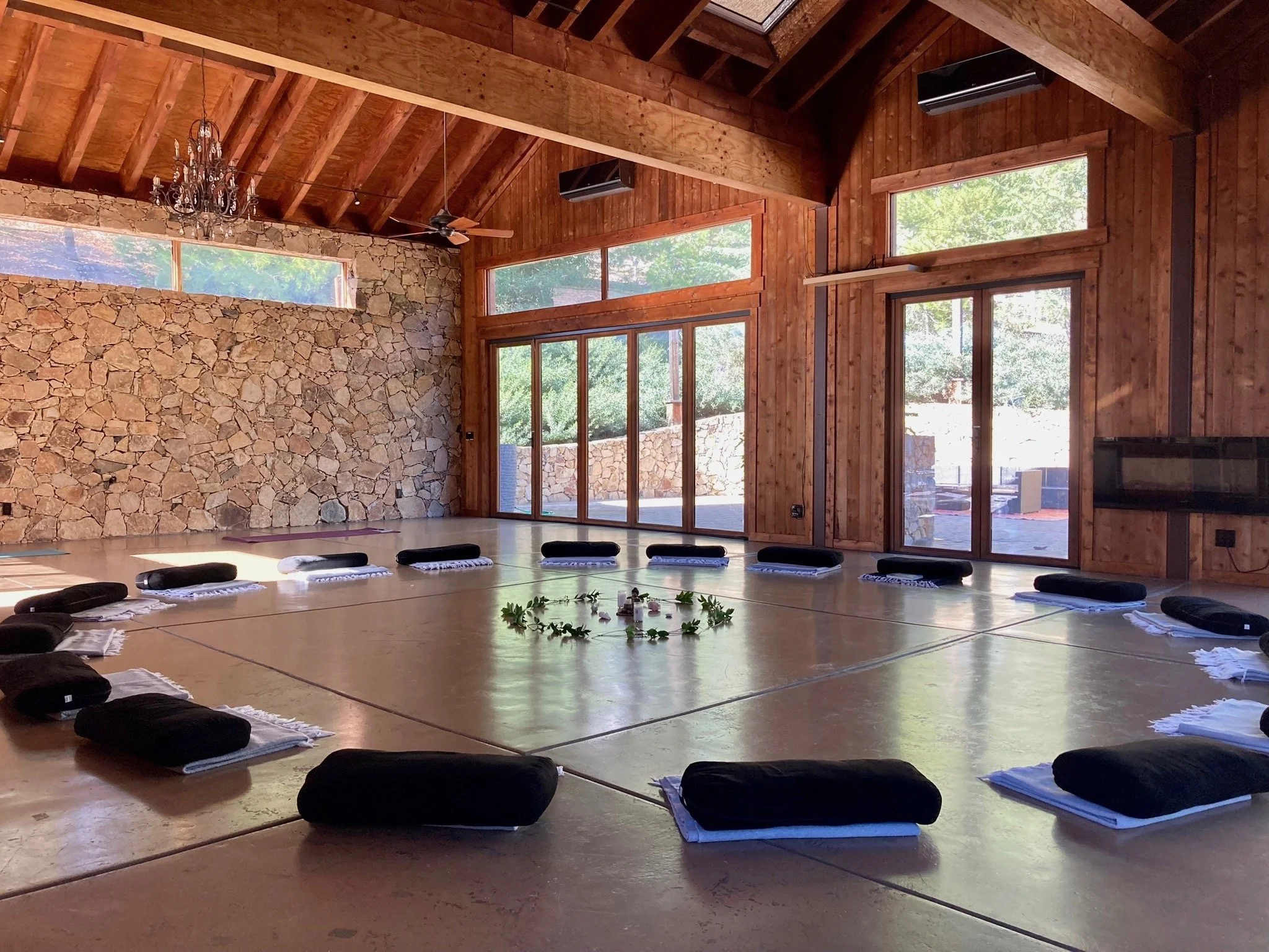 Rustic wood-and-stone meditation hall with black cushions arranged in a circle around a leafy centerpiece.