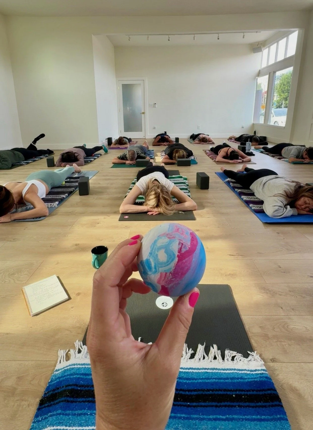 Yoga class participants resting on mats while an instructor holds a colorful lacrosse ball in a bright studio