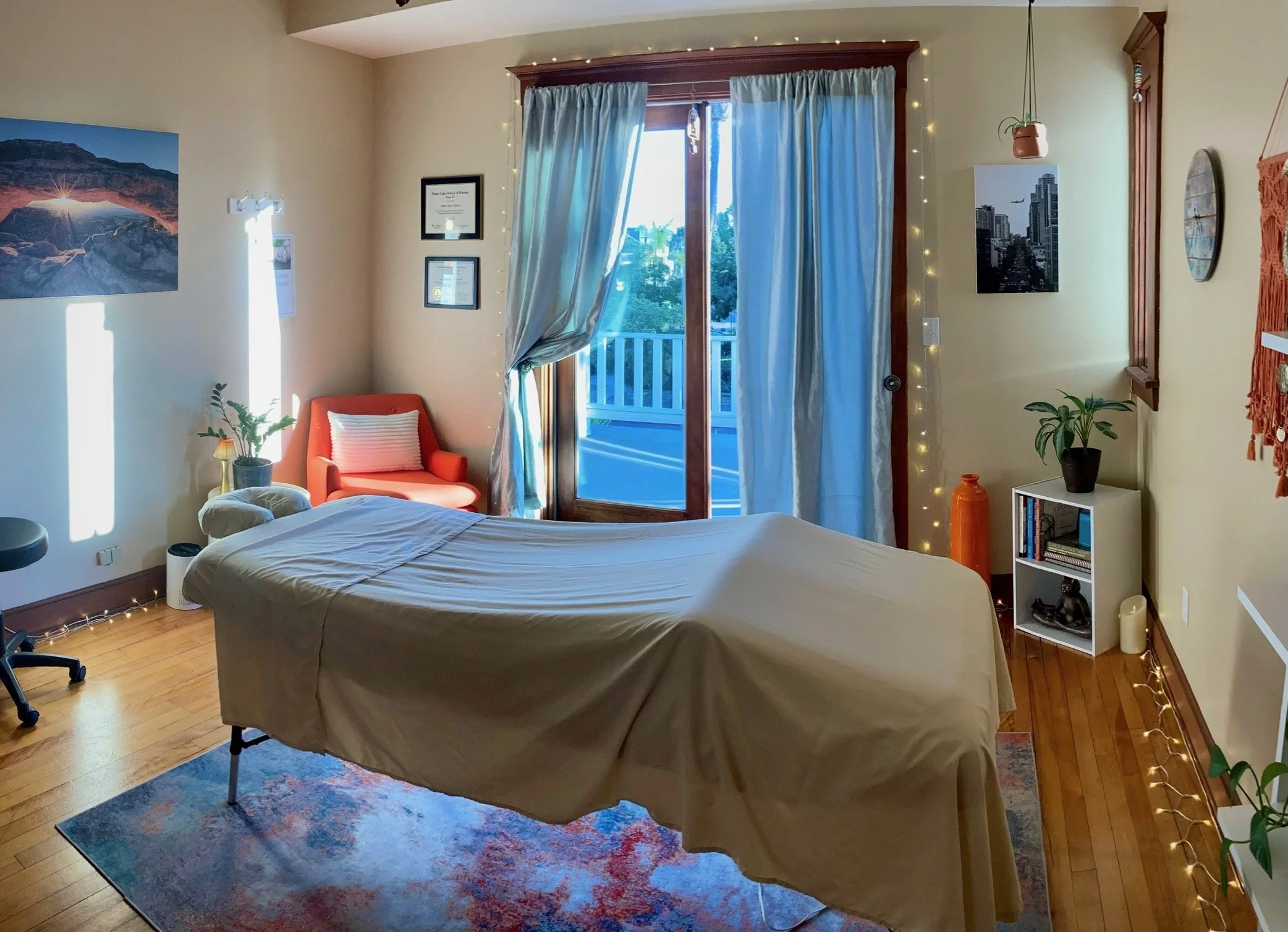 Cozy bodywork studio with massage table, blue curtains, balcony doors, orange armchair, plants, artwork, and string lights