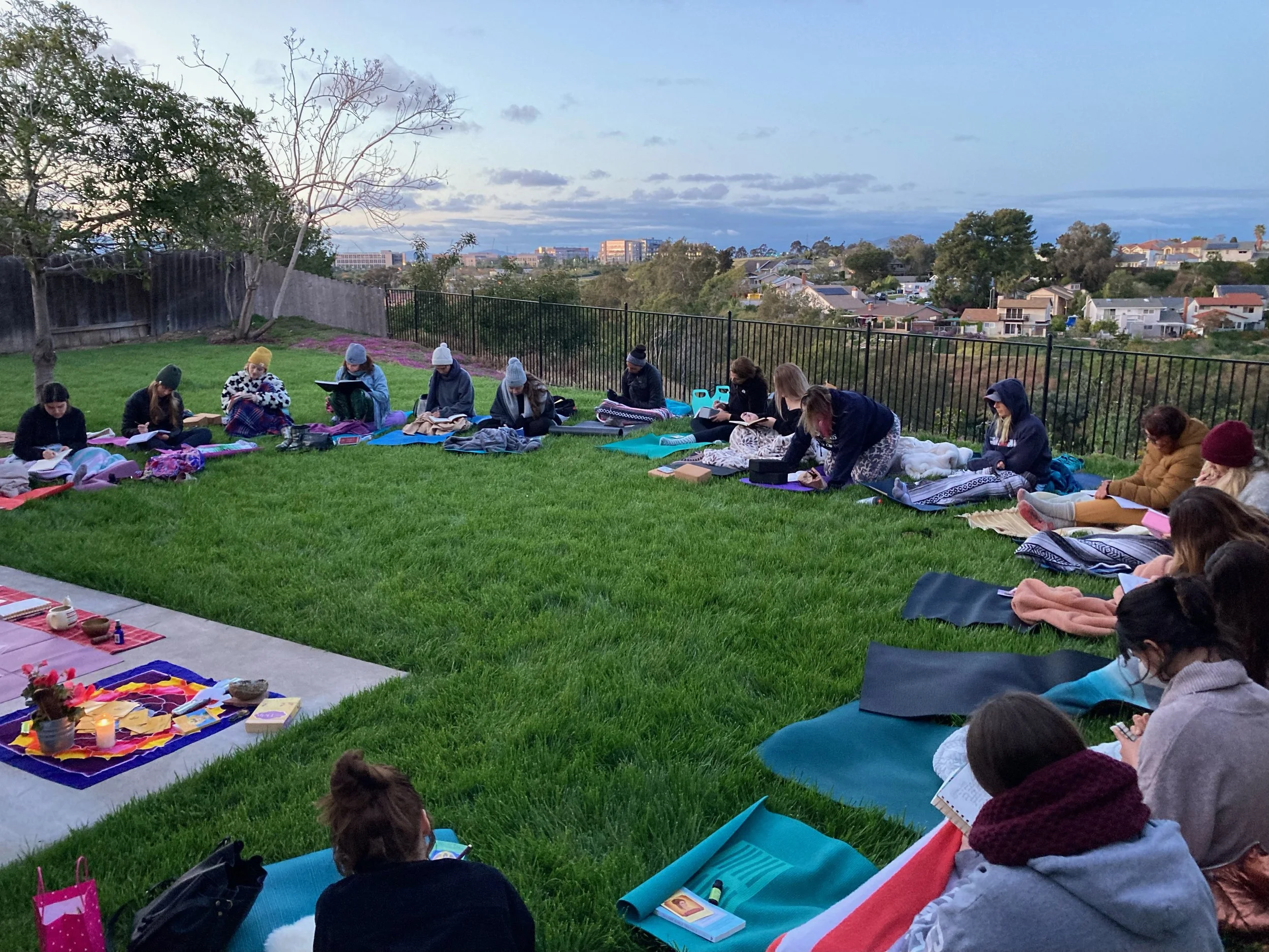 Group of people journaling on blankets and yoga mats in a grassy backyard overlooking a suburban neighborhood at sunset