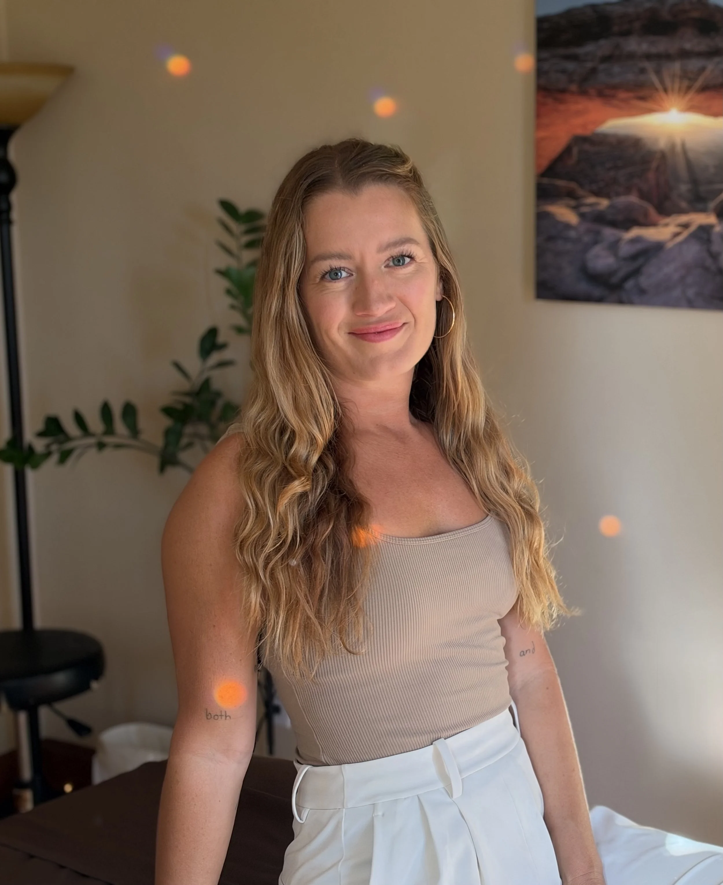Smiling woman with long blonde hair wearing a beige sleeveless top and white pants in a warmly lit bodywork studio