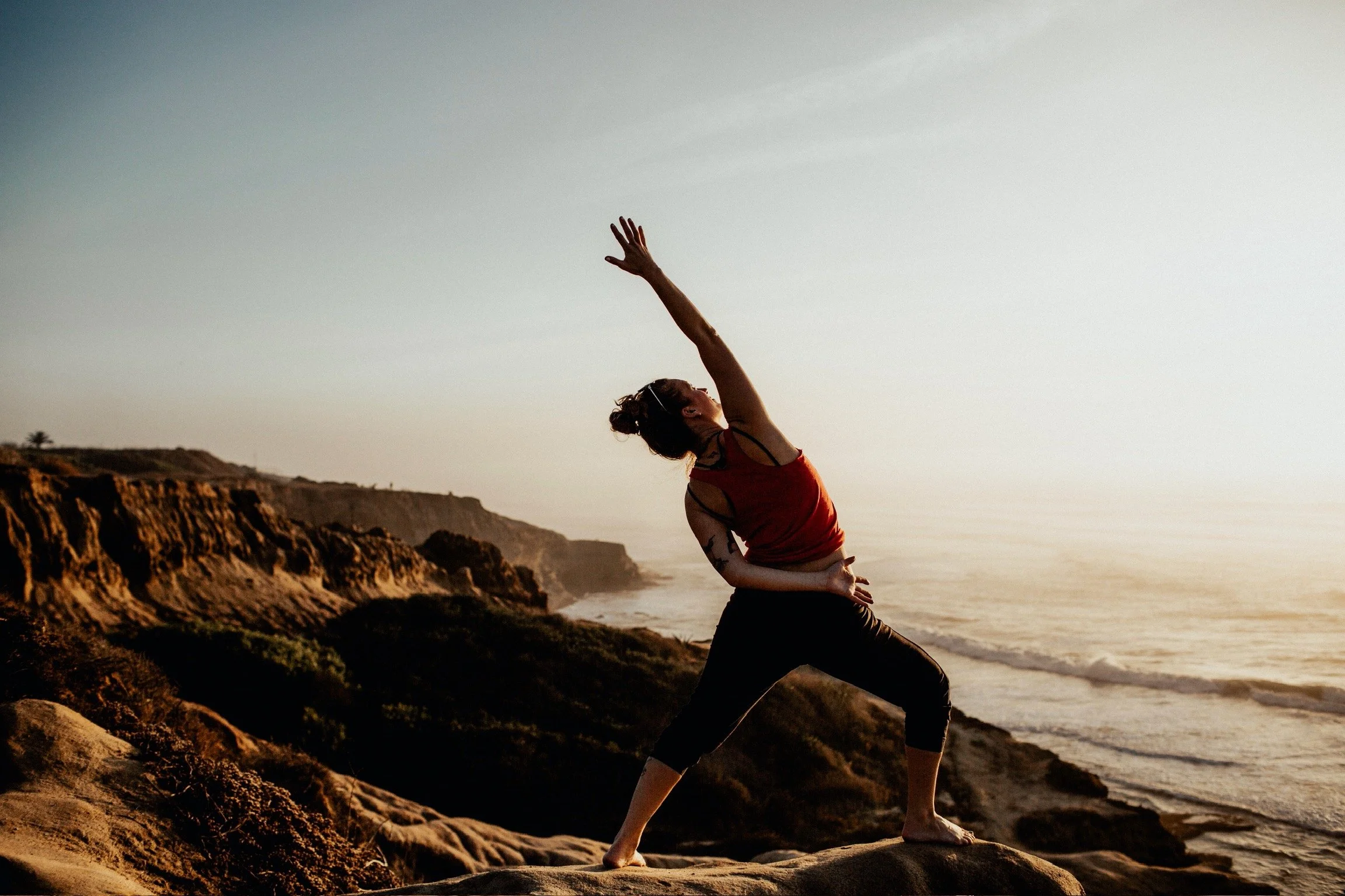 Woman practicing yoga on coastal rocks at sunset, with cliffs and ocean in the background