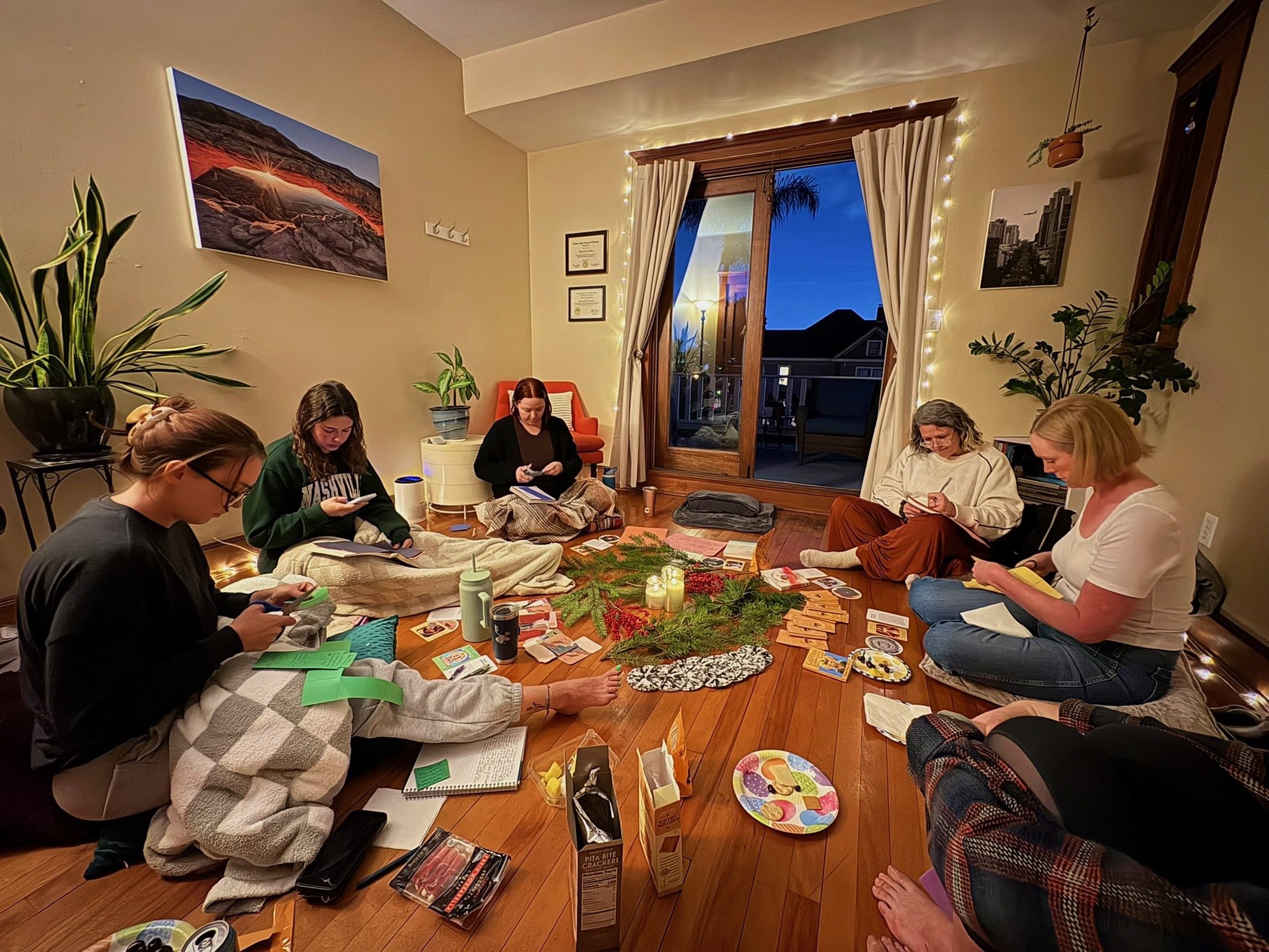 Group of women gathered indoors for a craft activity around a table decorated with greenery, candles, cards, snacks, and drinks.
