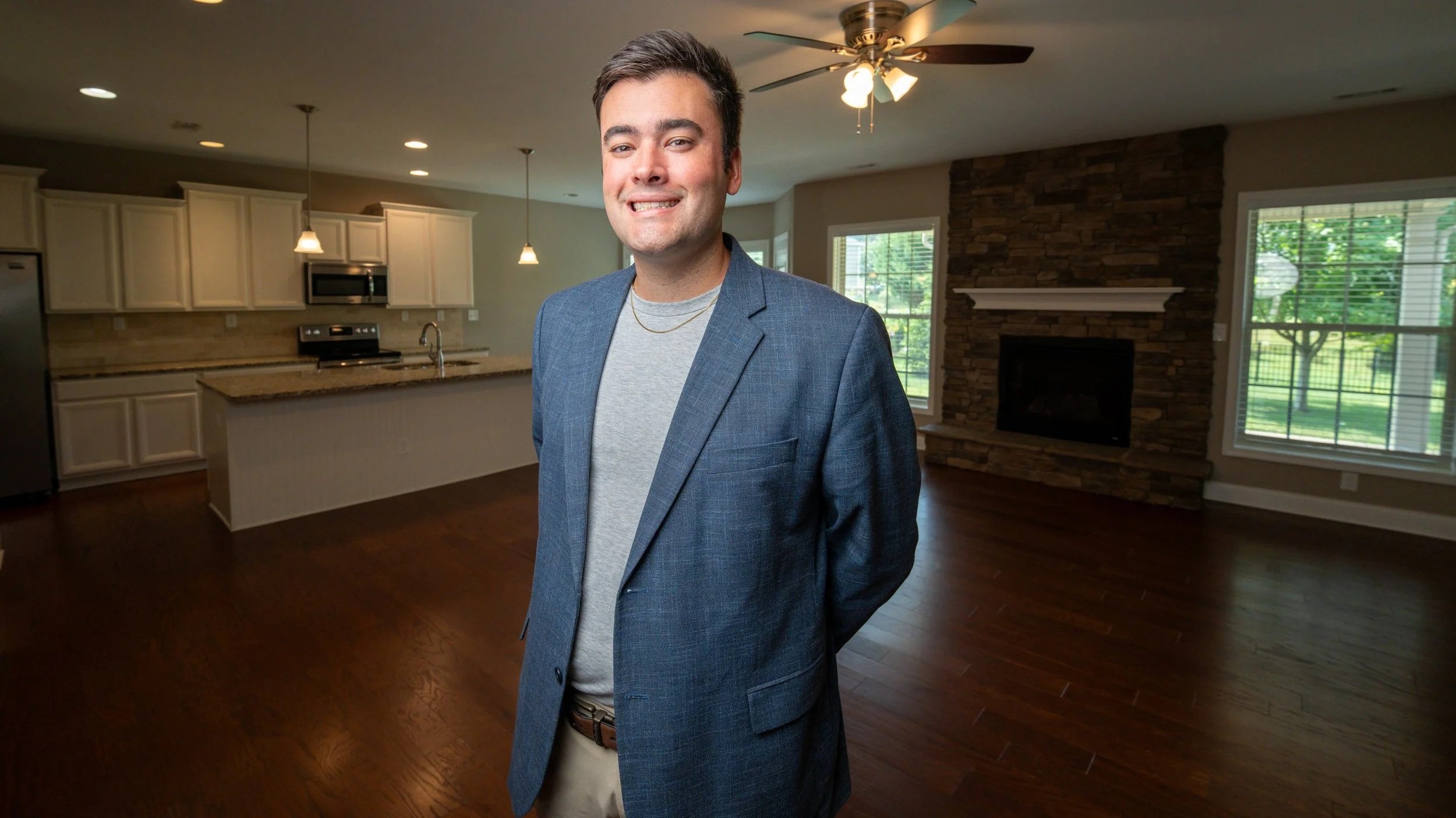 A man in a gray blazer and light gray shirt standing in a spacious living room with large windows, hardwood floors, and a modern fireplace.