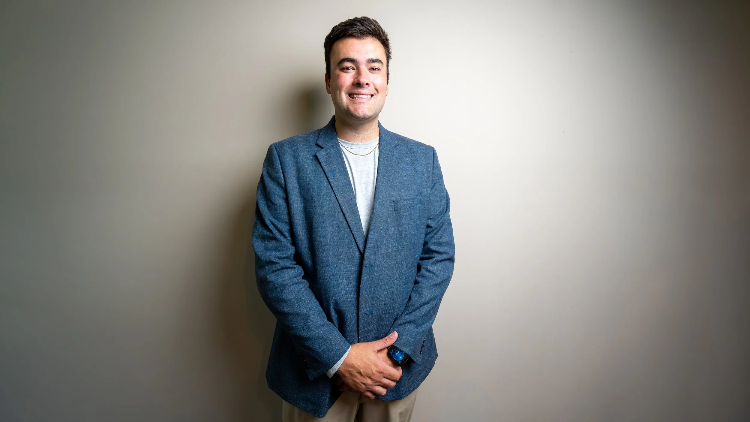 A young man standing in front of a plain wall, wearing a blue blazer, light gray t-shirt, and beige pants, smiling at the camera.