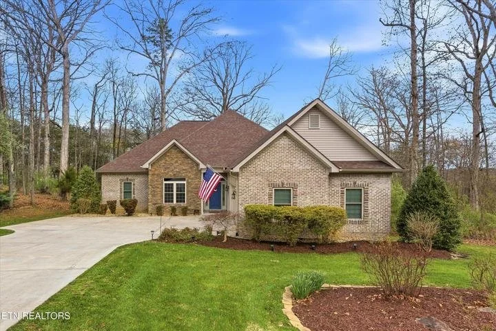 A new brick house with a brown roof and a driveway, surrounded by a landscaped yard with shrubs and trees, and an American flag by the front door.