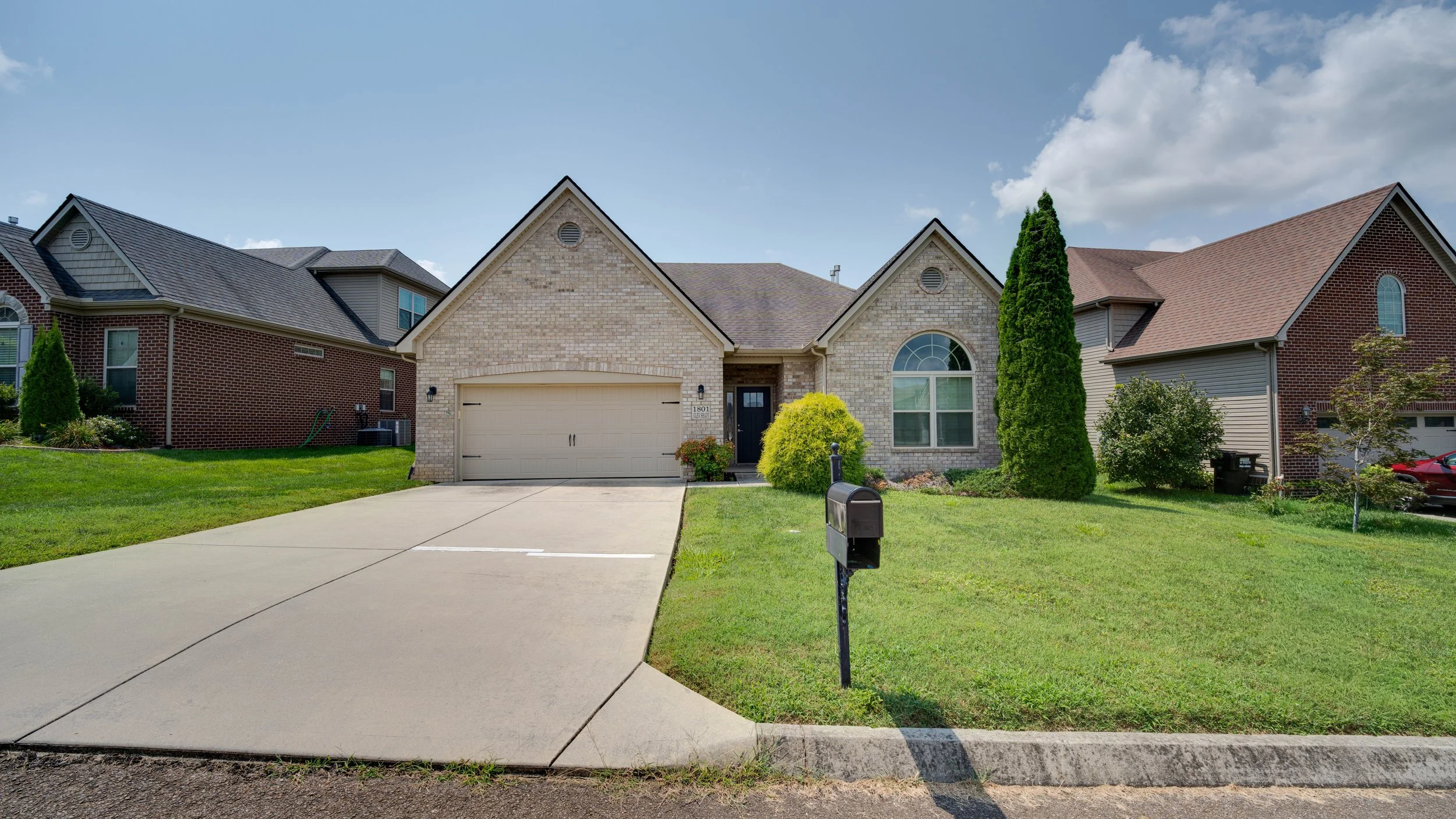 A suburban house with a beige garage door, brick and siding exterior, surrounded by a well-maintained green lawn, shrubs, and trees under a partly cloudy sky.