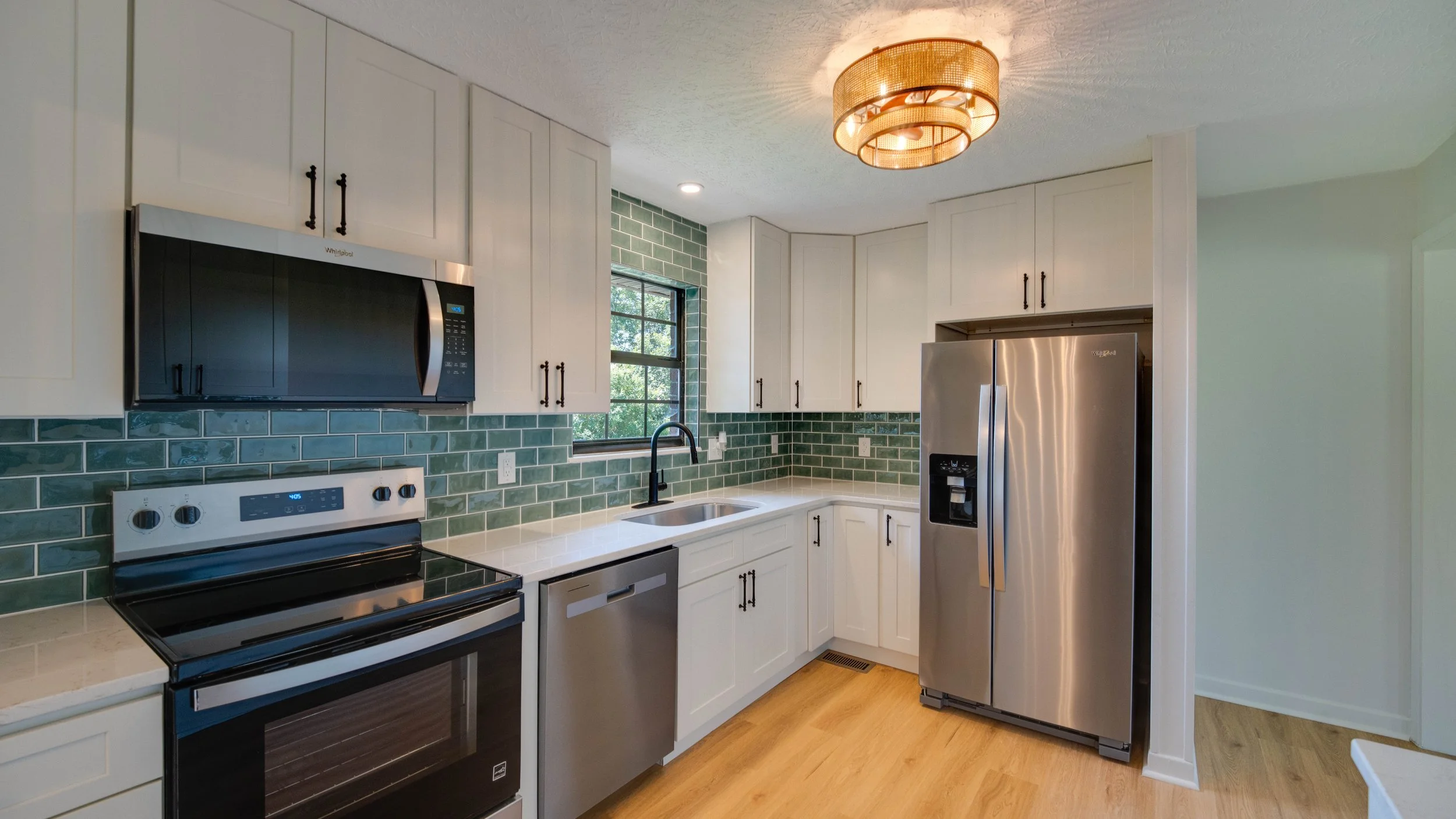 A modern kitchen with white cabinets, green tile backsplash, stainless steel appliances including a microwave, oven, dishwasher, and refrigerator, a black faucet, a window with black trim, and a ceiling light fixture.