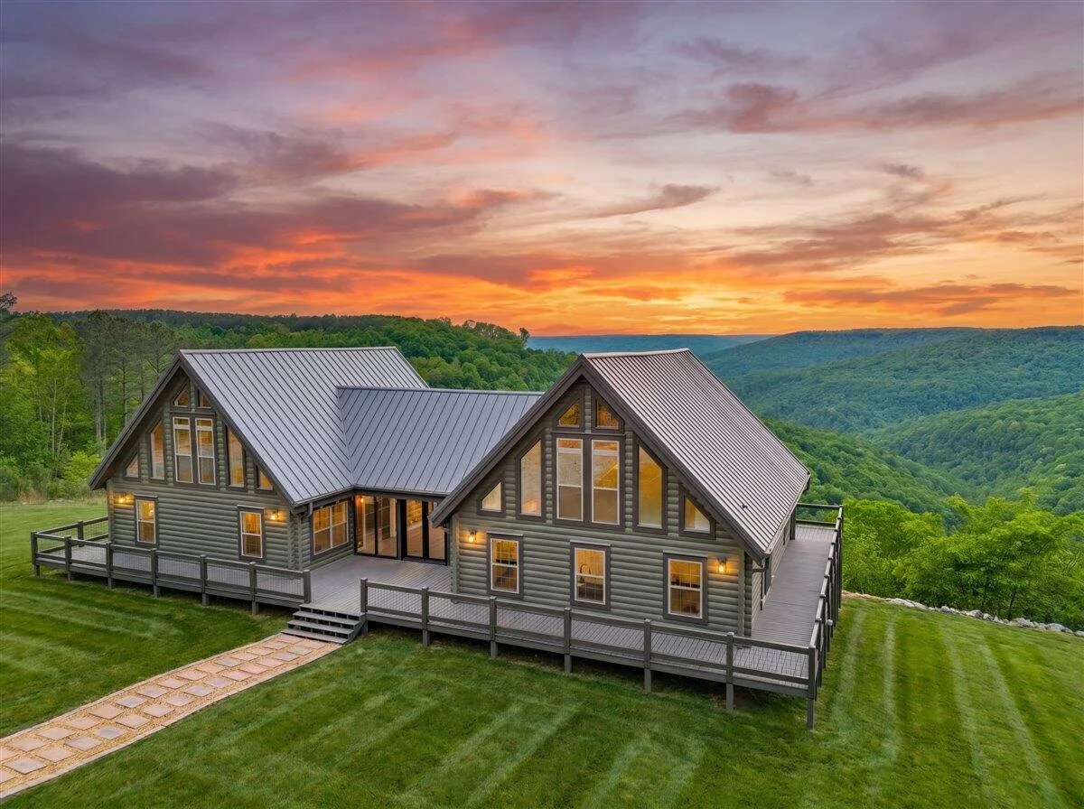 A modern house with large windows and a sloped metal roof, situated on a grassy hillside at sunset with a view of lush green mountains.