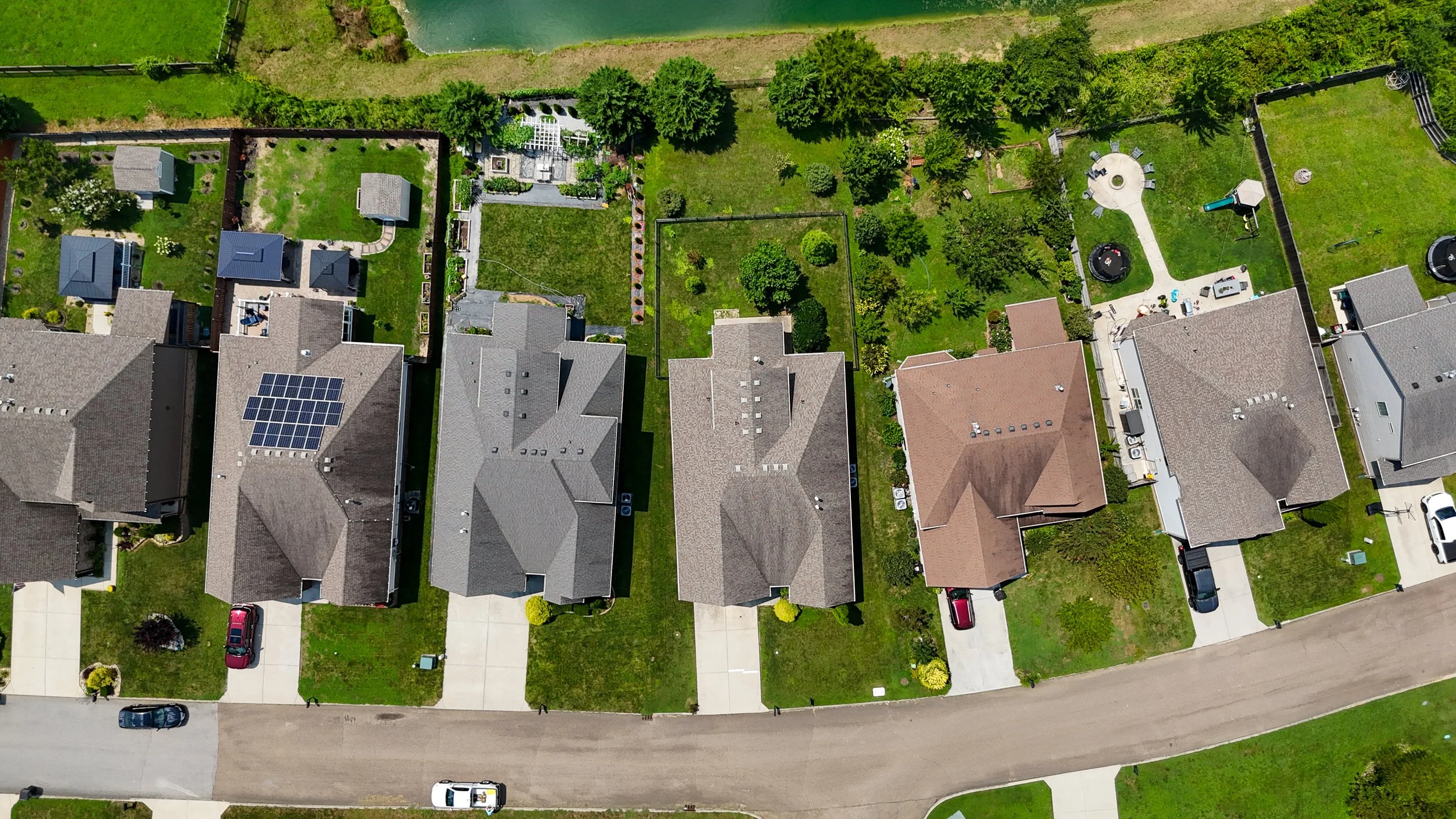 An aerial view of several suburban houses with backyards, some with solar panels on the roofs, neatly arranged along streets with green lawns, trees, and a small canal in the background.
