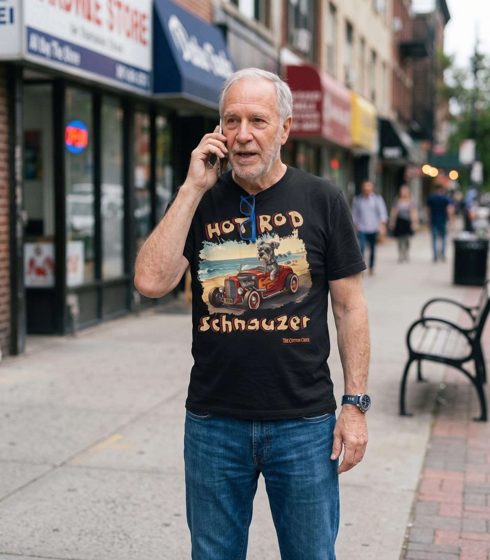 An older man with gray hair on his cell phone walking down a city street wearing a black unisex cotton t-shirt with graphics of a dog driving a hot rod car from the 50s. Design from The Cotton Crier.