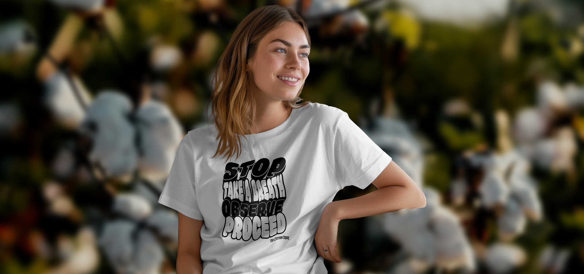 A young woman with brown hair, smiling and looking to the right, stands in front of a blurred background of cotton plants. She wears a white T-shirt with black text that shows the DBT Mindful Stacks design from The Cotton Crier. The Cotton Crier logo