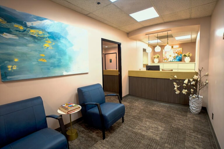 Waiting area of an office with two blue armchairs, a small side table with magazines, a large abstract painting on the wall, and a reception desk decorated with flowers in a vase.