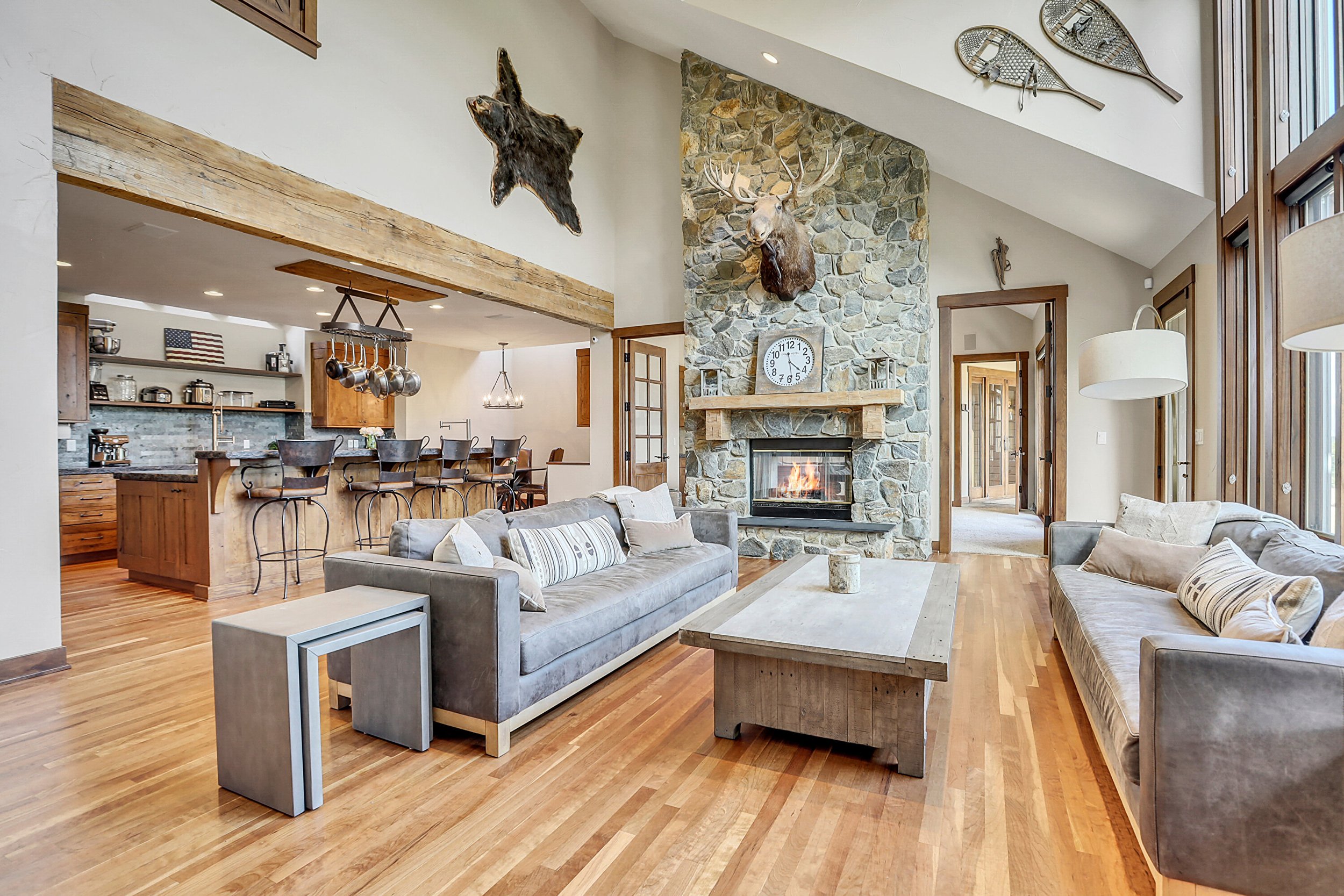 A spacious living room with a stone fireplace, mounted animal heads and vintage skis on the wall, wooden beams, and a kitchen in the background.