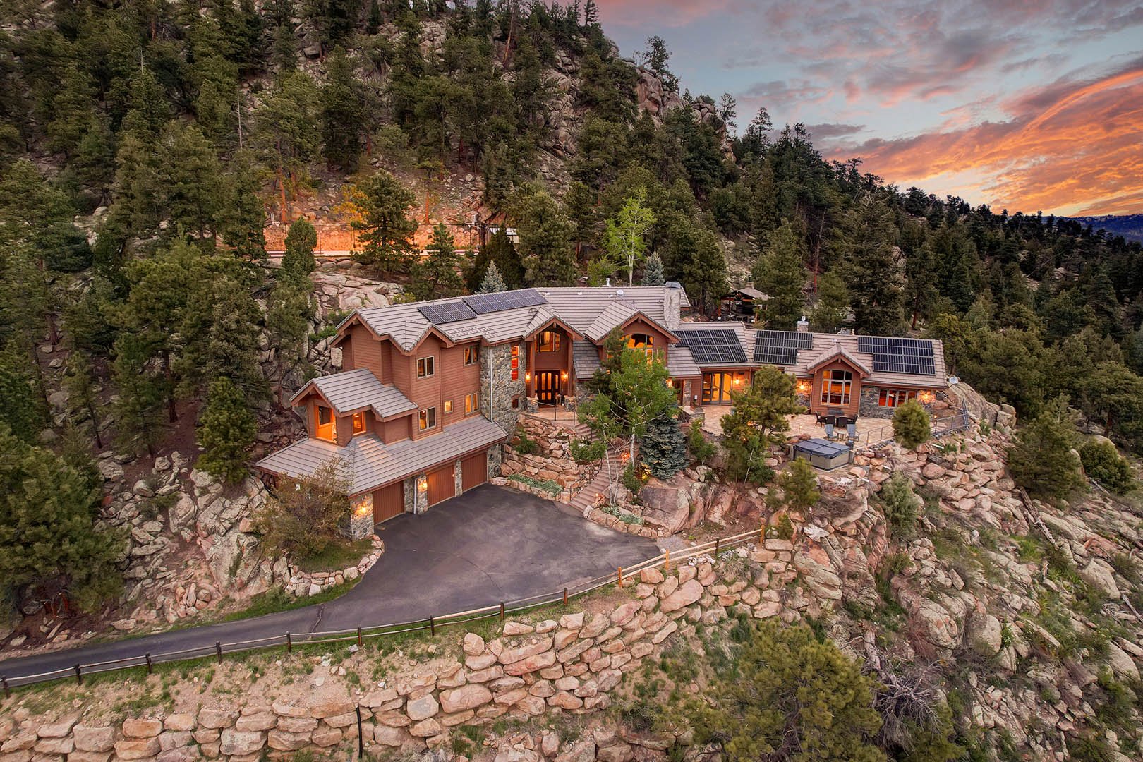 A large house built into a rocky mountainside surrounded by trees at sunset. The house has solar panels on the roof, multiple sections, and a driveway leading to a garage.