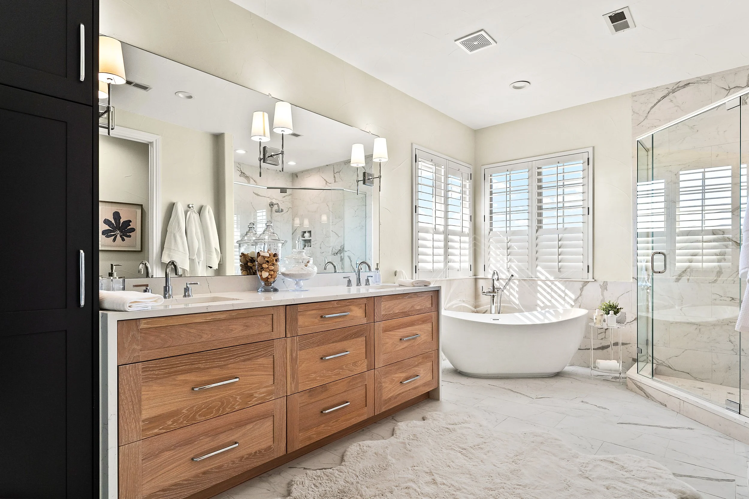 A modern bathroom with a large mirror, a wooden vanity with multiple drawers, a white bathtub, a glass shower enclosure, and windows with white plantation shutters, all decorated in a neutral color palette.