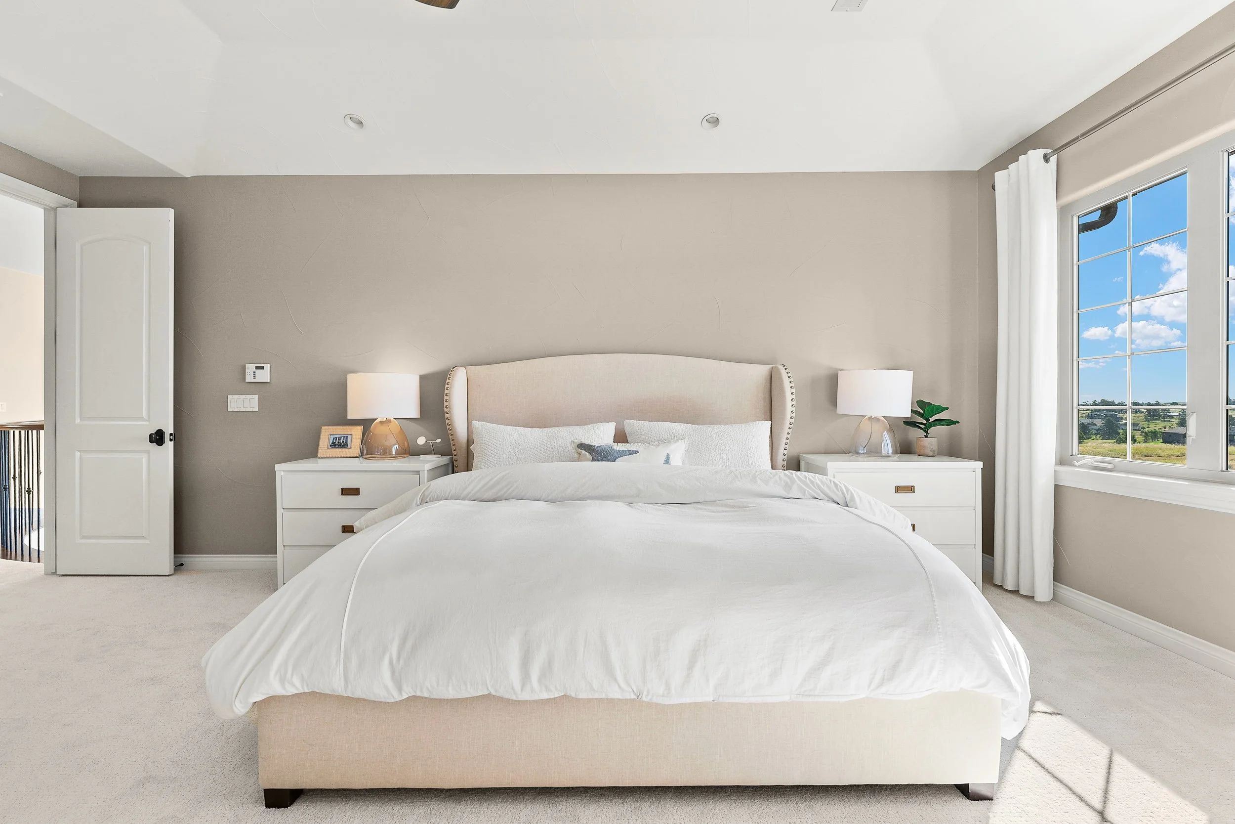 A bedroom with a large bed, white linens, and a beige upholstered headboard. Two white nightstands with lamps and decorative items sit on either side of the bed. A window with white curtains and a view of blue sky and green landscape is on the right side.