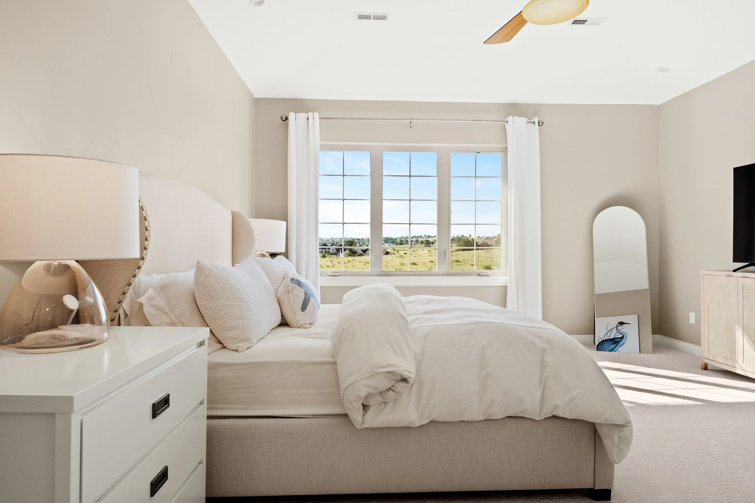 A bedroom with a white bed, white nightstand, and window with white curtains showing a view of open land and houses. The ceiling has a ceiling fan with wooden blades, and a small framed bird artwork is leaning against the wall near the dresser.