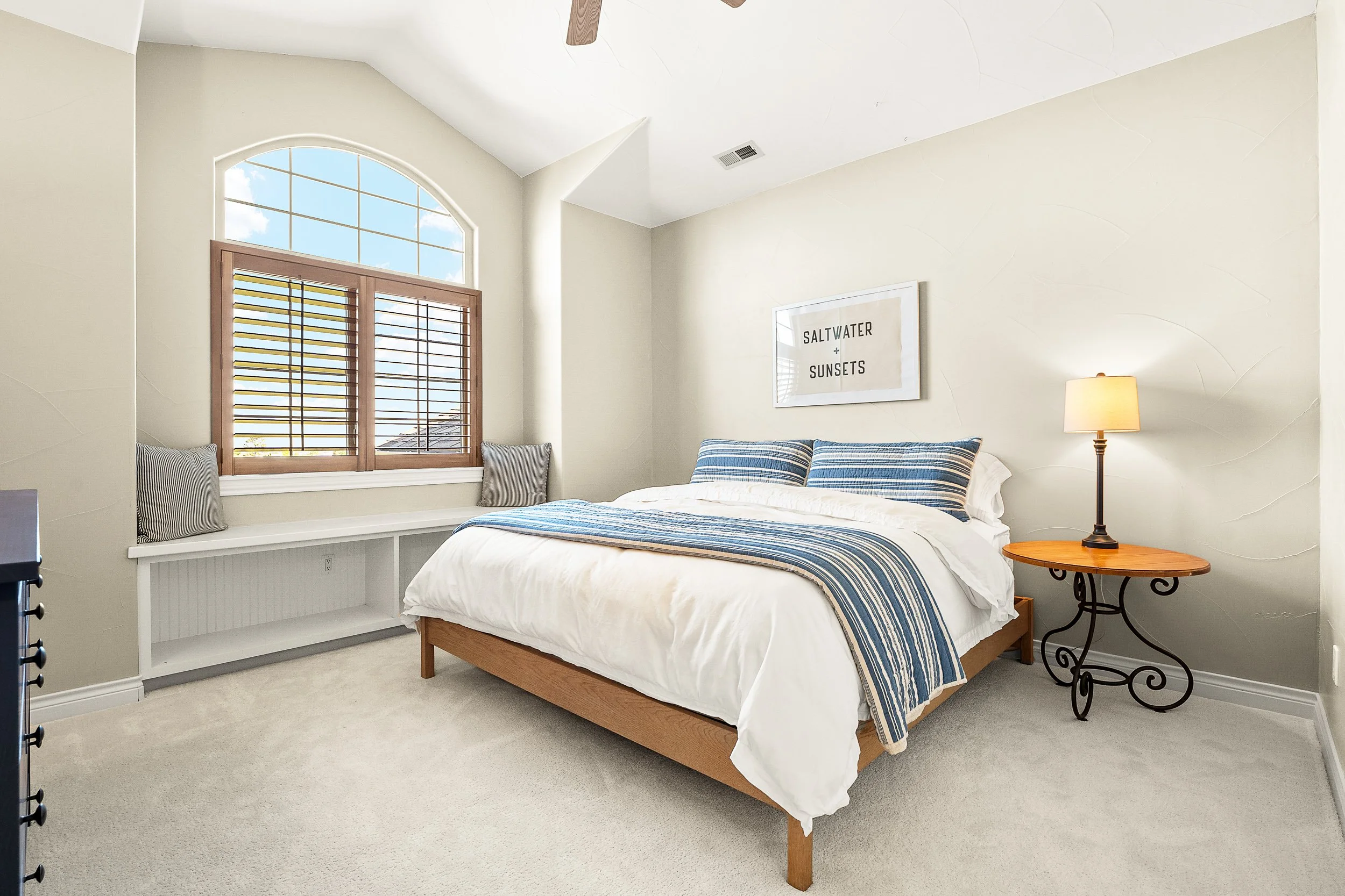 A bedroom with a bed featuring blue and white striped bedding, a wooden side table with a lamp, a framed wall art with the text 'Saltwater + Sunsets', a window with wooden shutters, and a chair with a cushion near the window.