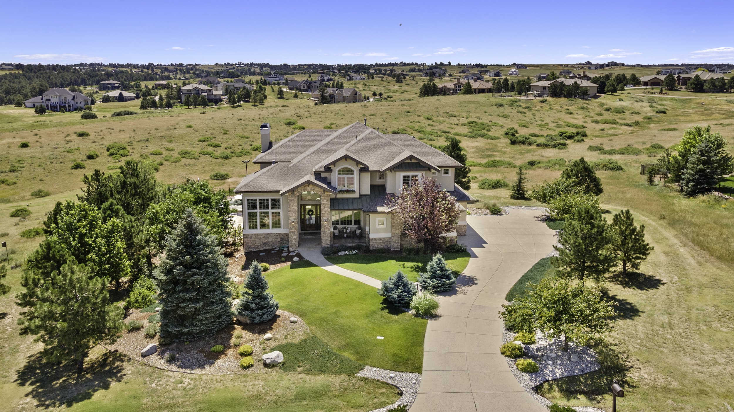 Aerial view of a large house with front lawn, trees, and a driveway, surrounded by open land and neighboring houses under a clear blue sky.