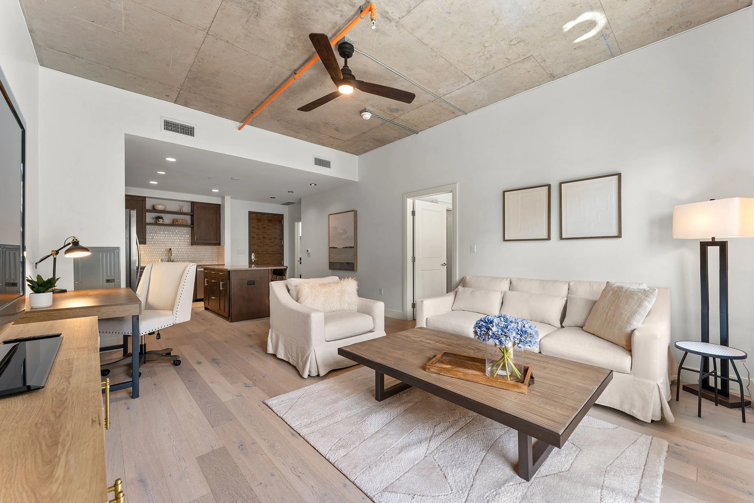 Living room with white sofa, beige armchair with fur throw, wooden coffee table with blue flowers, modern dining table with chairs, ceiling fan, artwork on walls, open kitchen in background, mixed ceiling materials, wooden floors, lamp, and minimal decor.