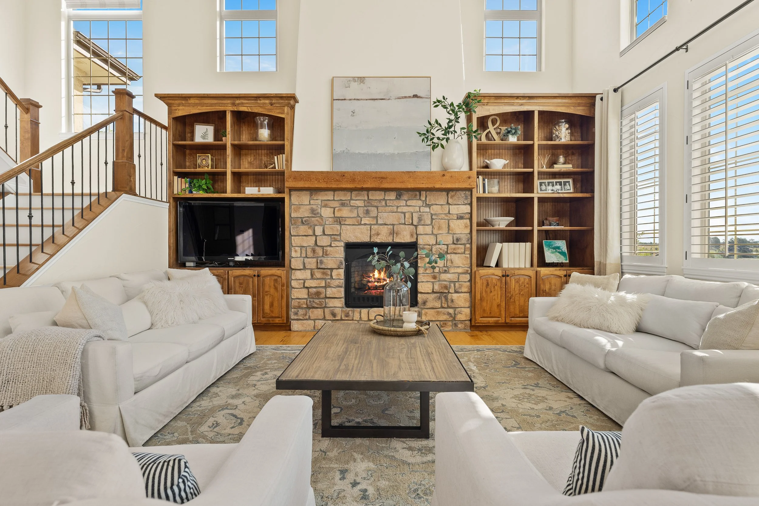 Living room with white sofas, a central wooden coffee table, a brick fireplace, built-in wooden shelves, large windows with white shutters, and decorative items on the shelves and mantel.