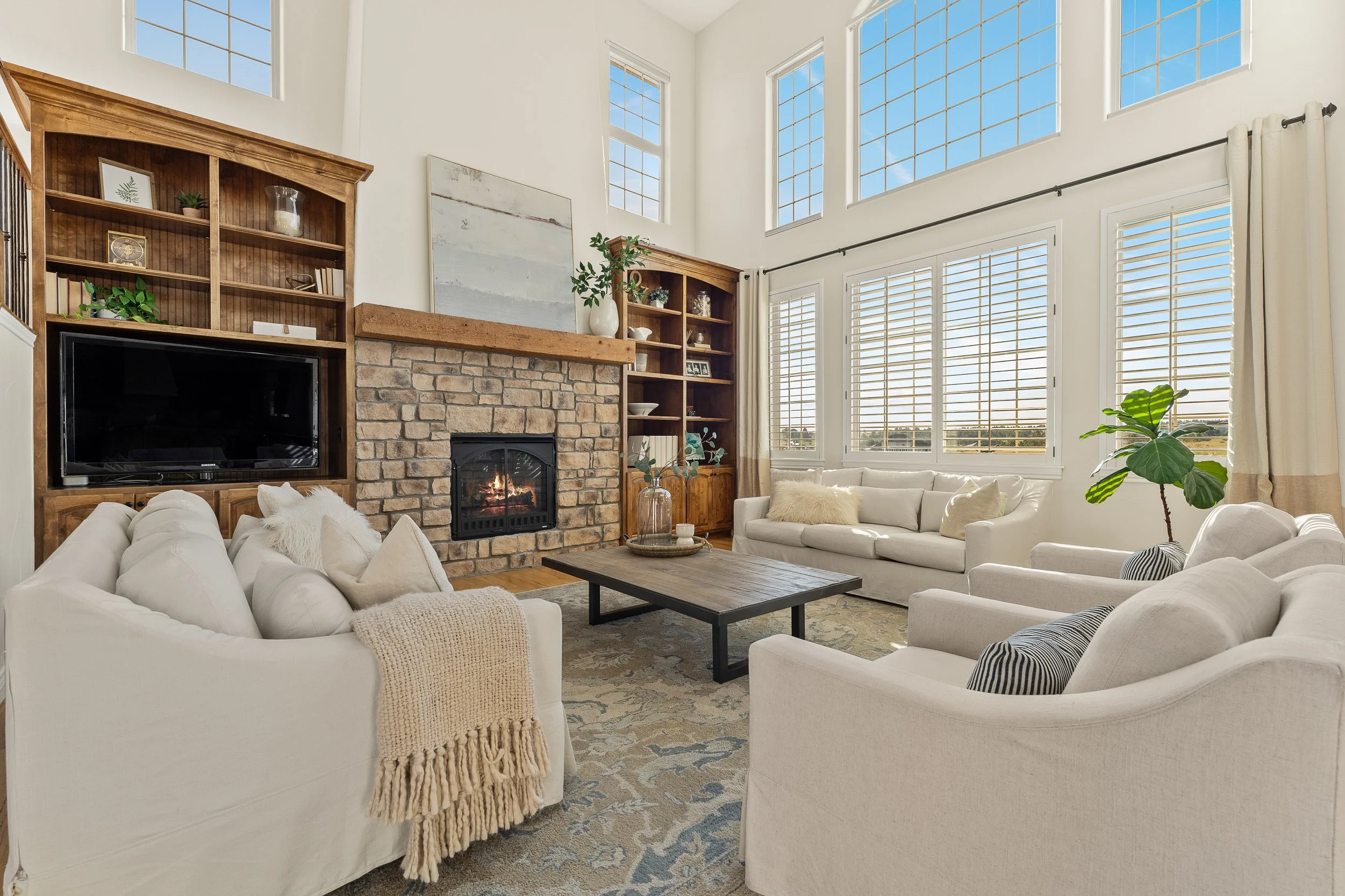 Living room with white sofas, a brick fireplace, wooden bookshelves, large windows with blinds, and houseplants, creating a bright, cozy space.
