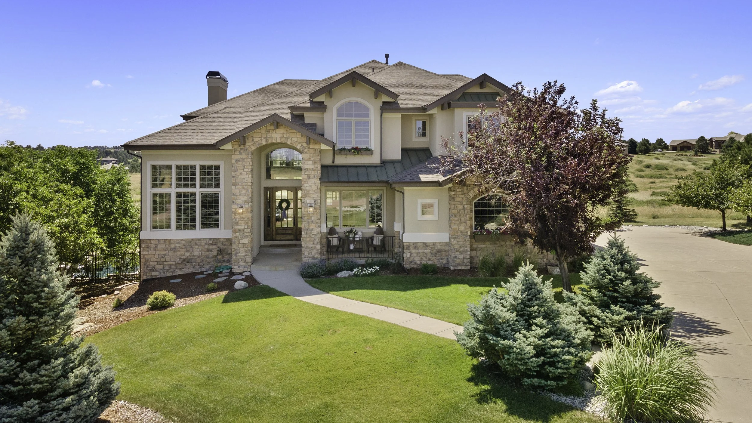 A two-story house with a brick and stucco exterior, large front windows, and a small porch with outdoor seating. The yard has green grass, landscaped bushes, and trees, with a clear blue sky overhead.