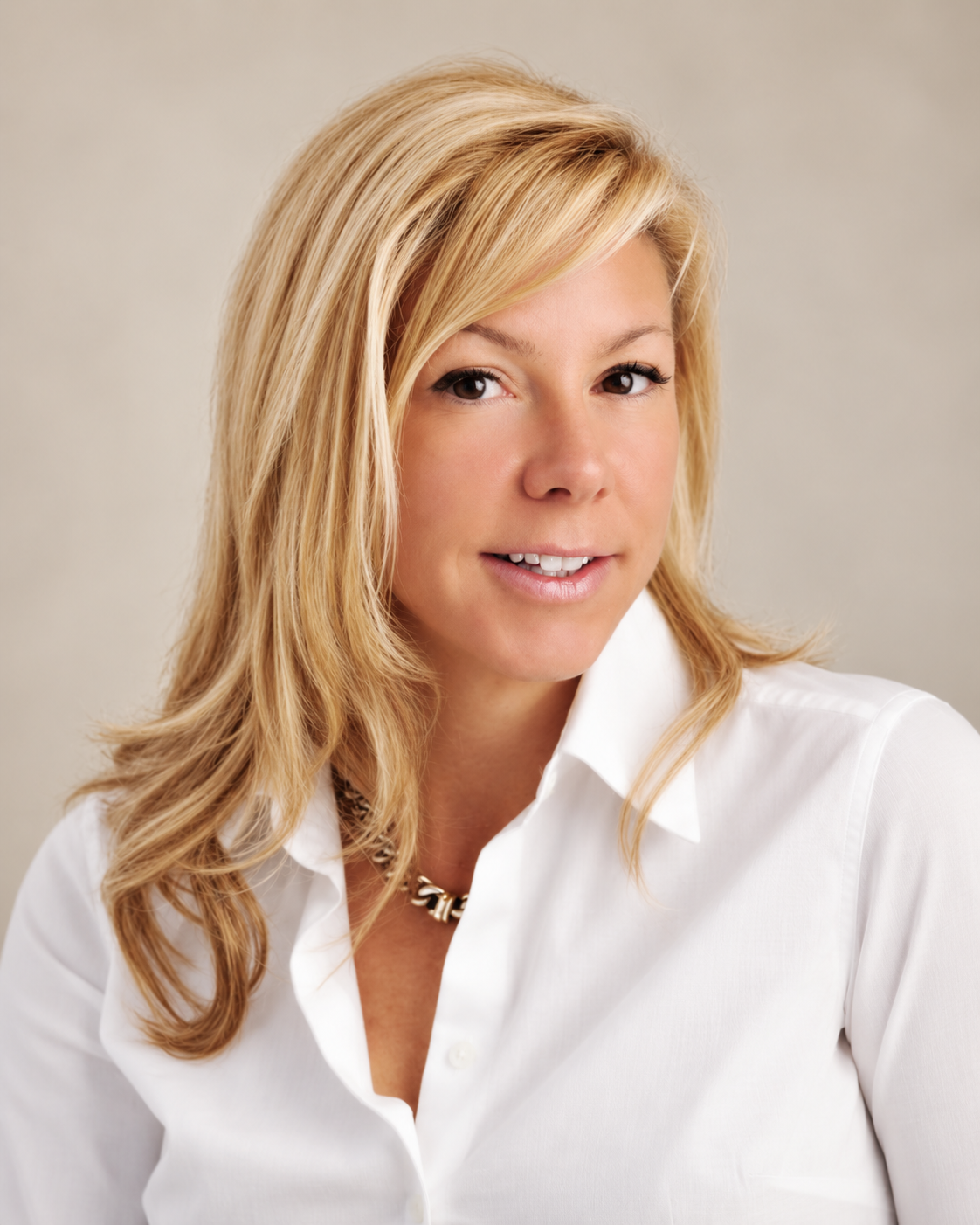 A woman with blonde hair wearing a white shirt and a gold chain necklace, smiling at the camera.