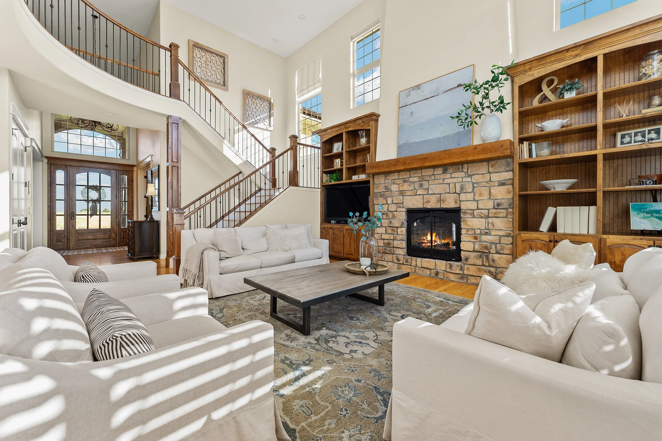Living room with white sofas, a fireplace, wooden shelves, and large windows letting in sunlight.
