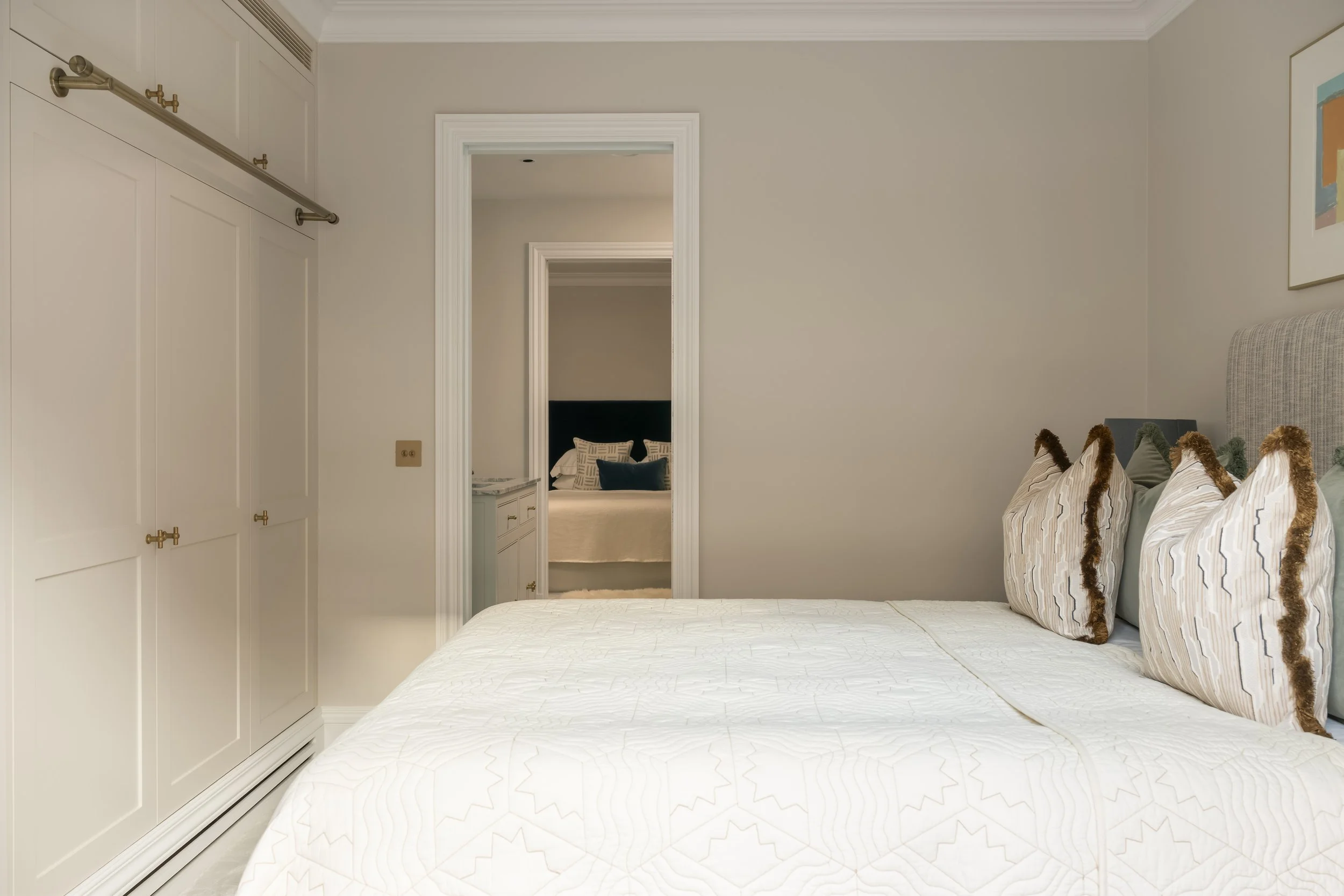 A bedroom with a white bed, striped pillows with fur accents, a built-in wardrobe, and a doorway leading to another room with a bed and a nightstand.