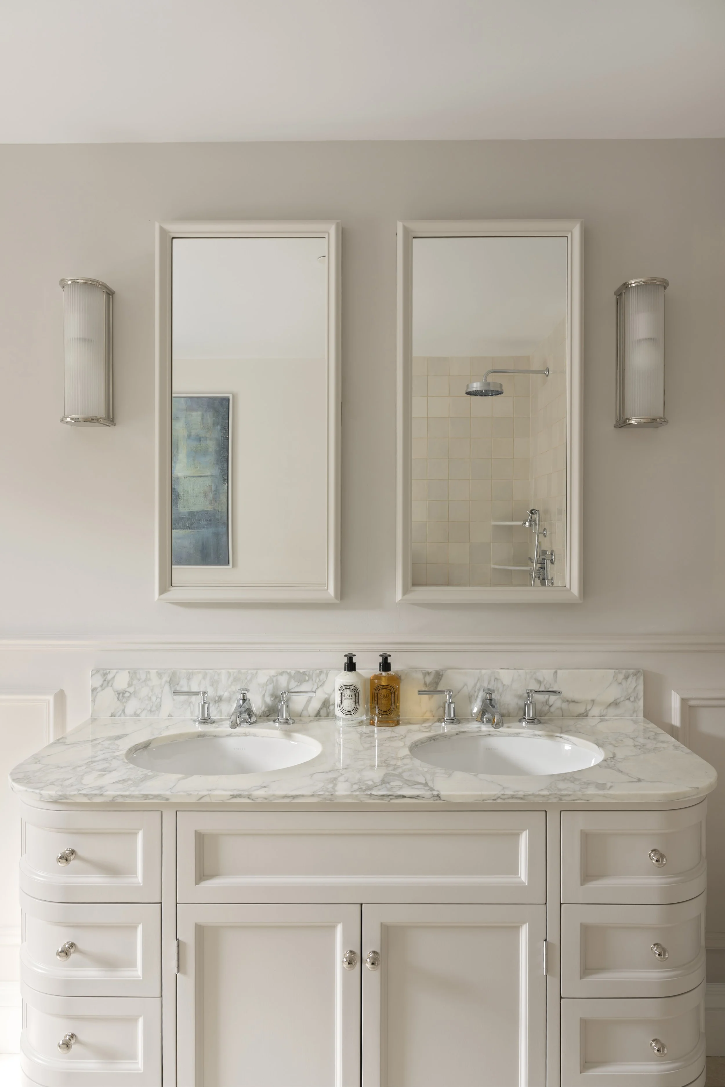 Bathroom with a double sink marble countertop, white cabinets, two wall-mounted light fixtures, and a mirror reflecting a shower area with beige tiled walls and a showerhead.