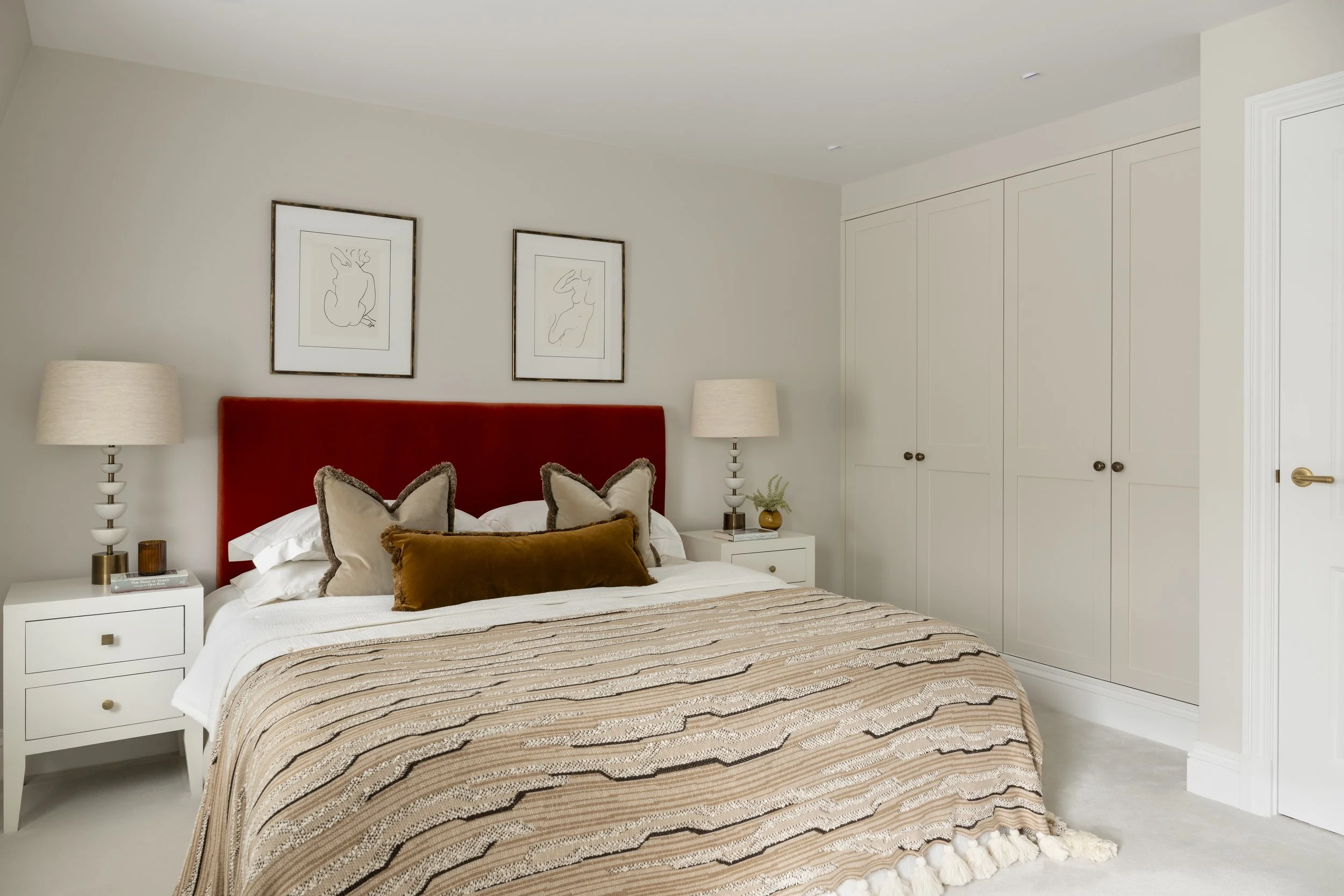 A cozy bedroom with a bed featuring a red velvet headboard, decorative pillows, and a striped bedspread. There are two white nightstands with matching lamps on each side of the bed. Above the bed are two framed line art illustrations of abstract human forms. The room has white walls, built-in closet doors, and a door on the right side.