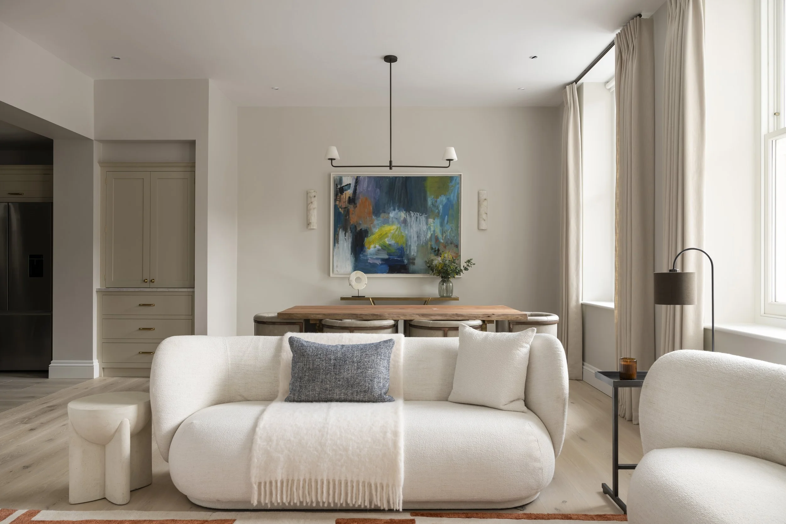 Living room with white sofa, decorative pillows, abstract wall art, and large windows with cream curtains.
