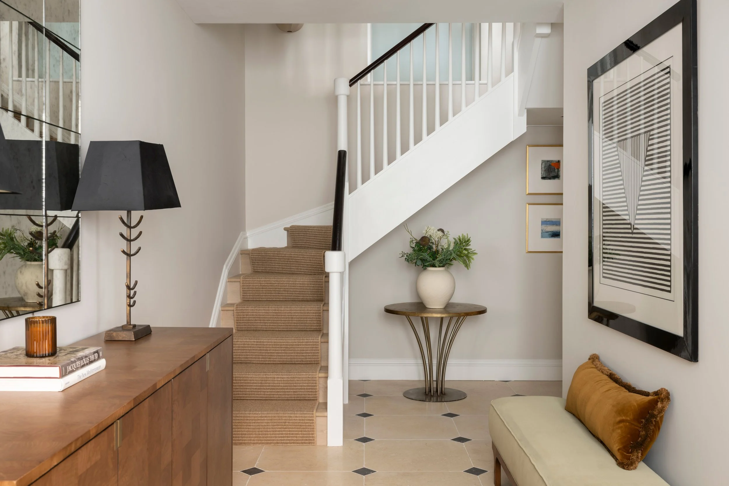 Interior view of a home entryway and living area featuring staircase with beige carpet, white walls, black framed artwork, a small round table with a floral arrangement, a wooden console table with books and a lamp, and a mustard-colored bench with a brown pillow.