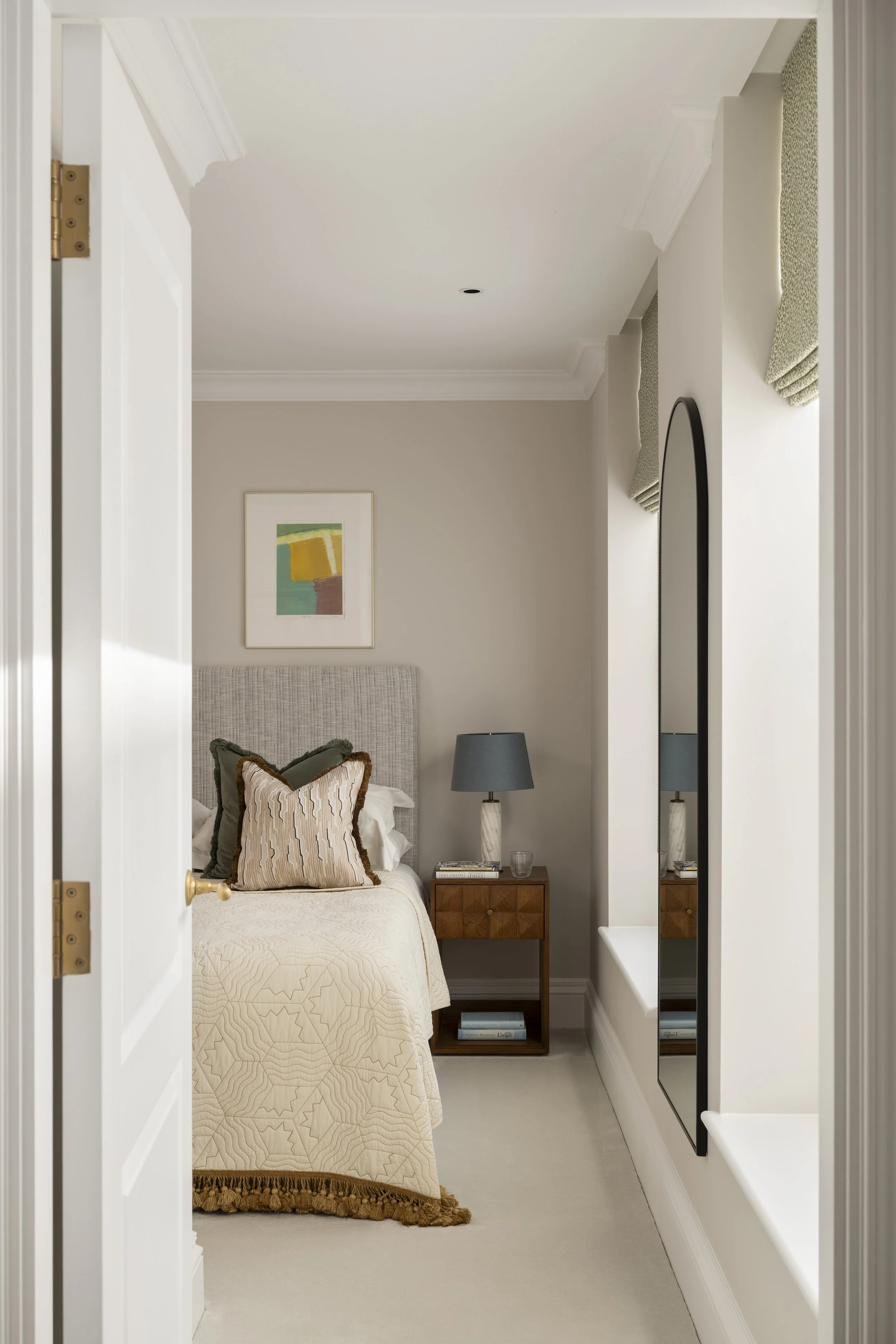 A cozy bedroom with a bed, patterned pillows, and a beige quilted bedspread. There's a wooden nightstand with books and a gray table lamp. A framed abstract artwork hangs on the wall above the bed, and windows with green fabric valances are on the right. A tall mirror reflects part of the room, and a white wall with crown molding surrounds the space.