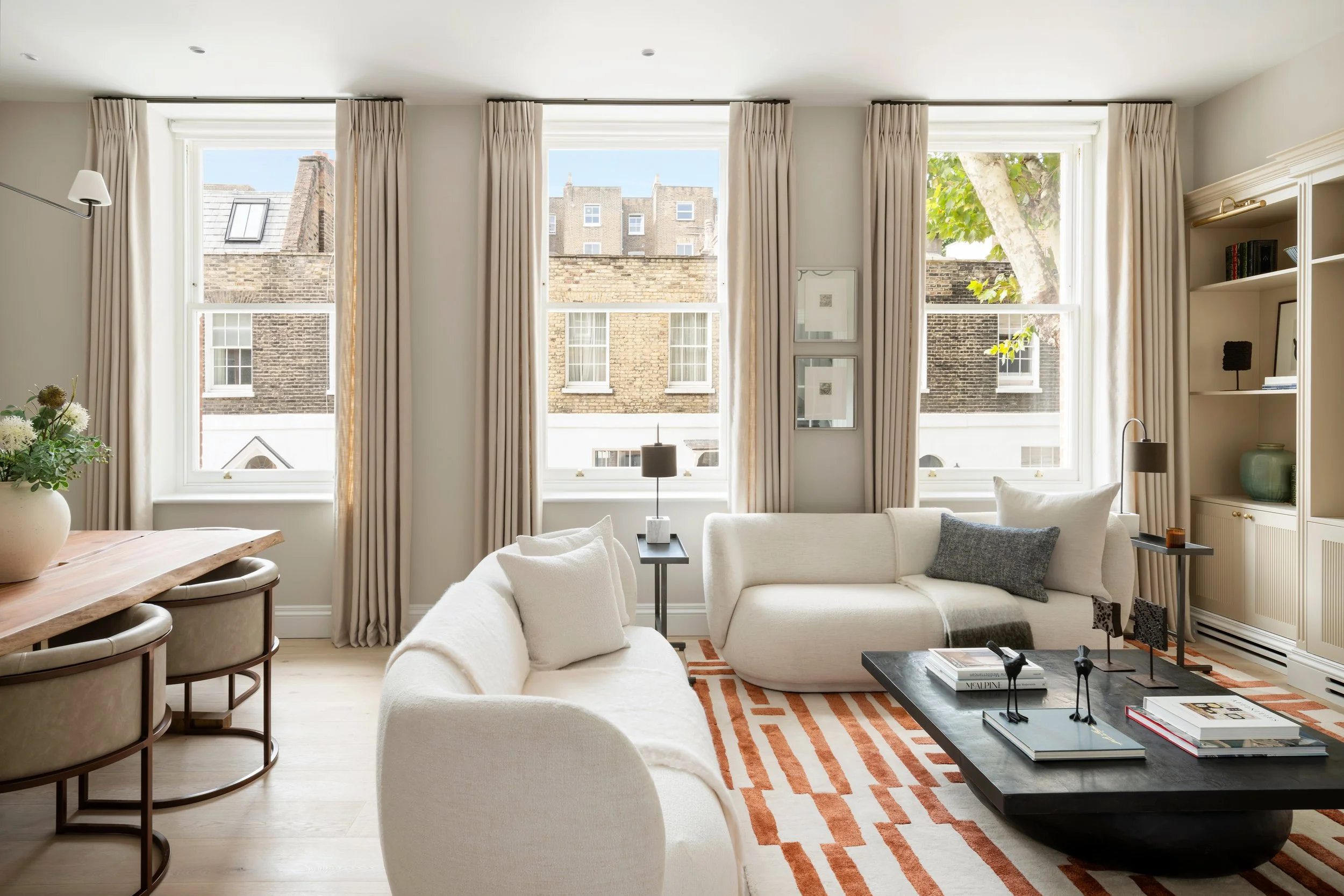 Living room with large windows, beige curtains, a white sofa, a coffee table, and decorative items, with neighboring buildings visible outside.