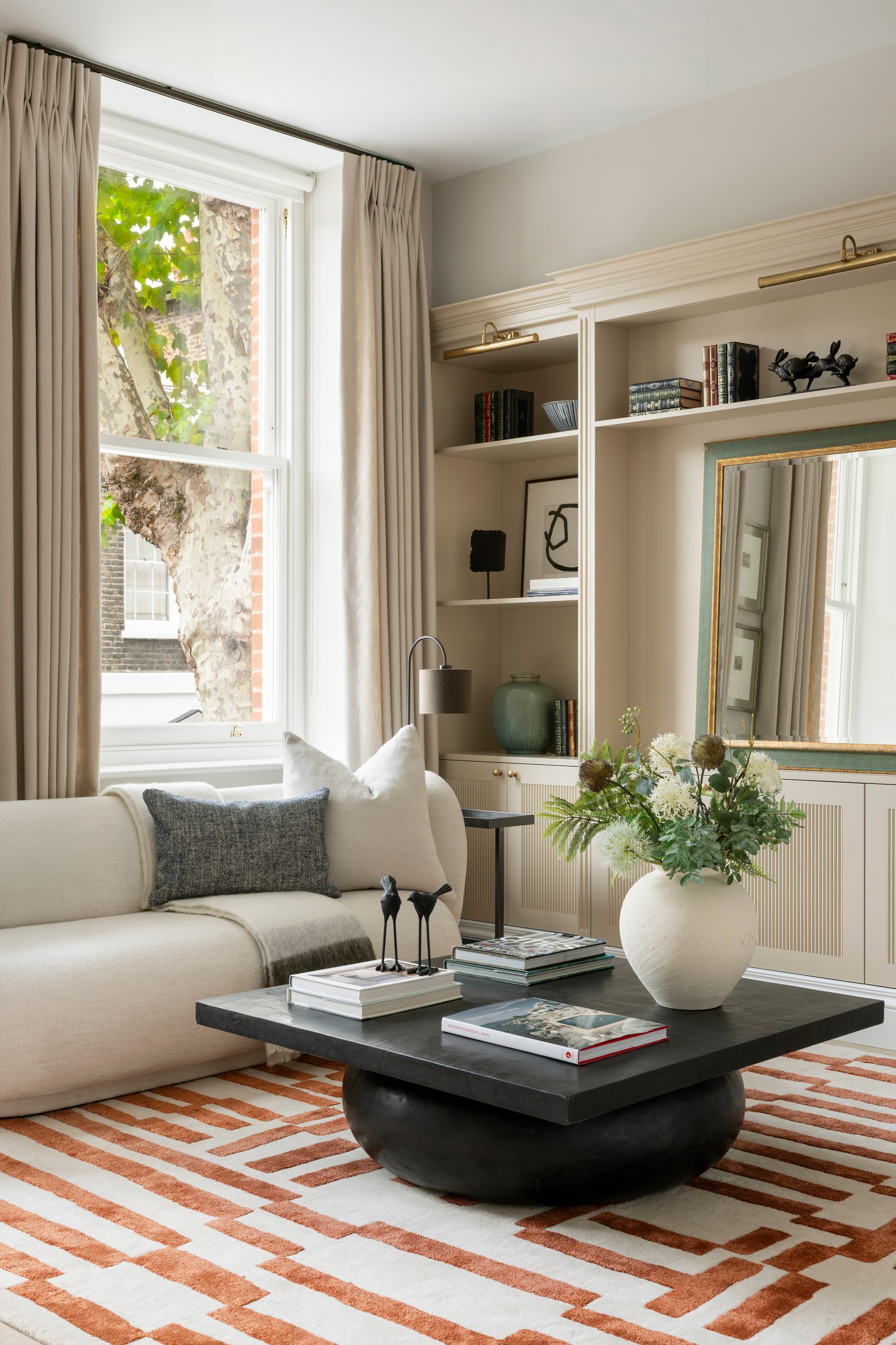 Living room with large window, beige sofa, black coffee table with books and decorative objects, white built-in shelves with books and art, large ceramic vase with flowers, striped rug, and framed mirror.