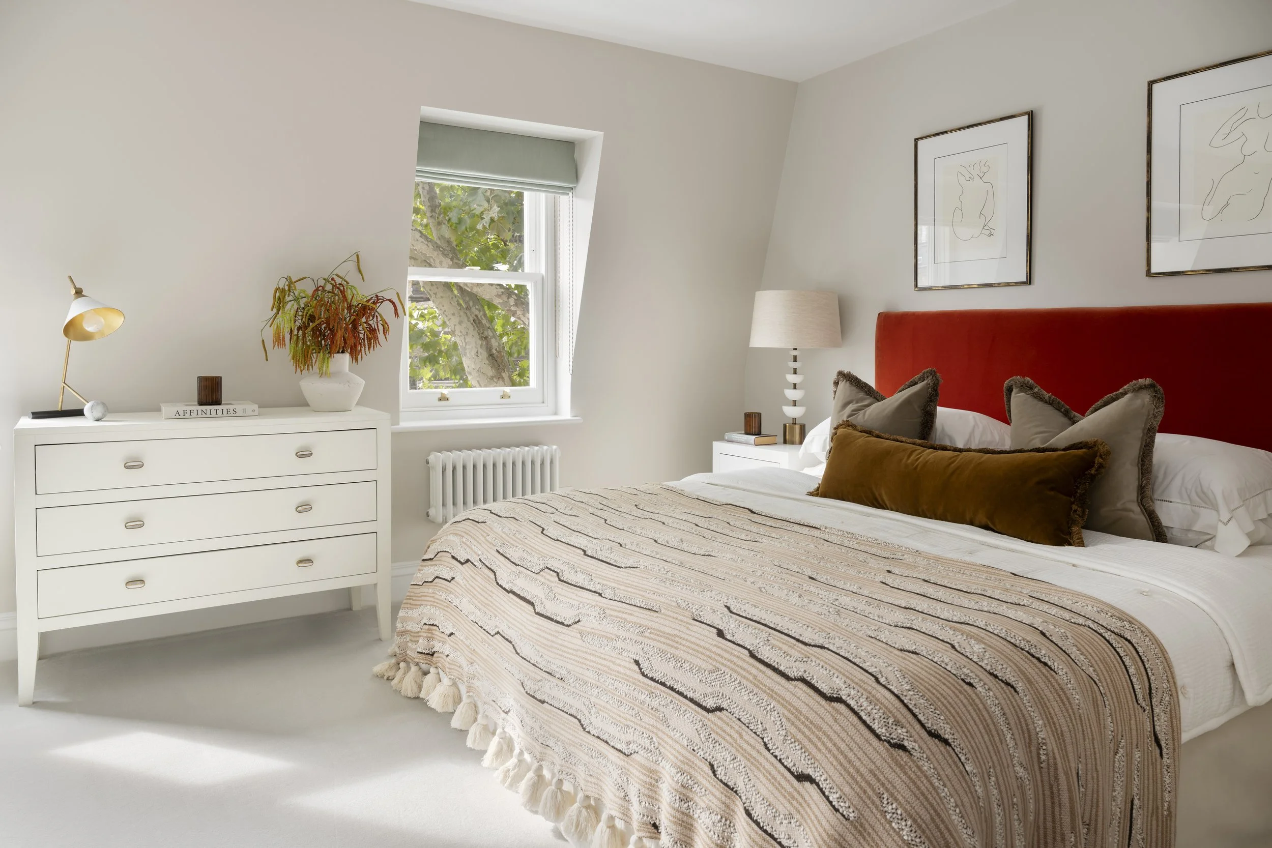 A bedroom with a white dresser, a window with trees outside, and a bed with an orange headboard and decorative pillows. There are framed line art drawings on the wall above the bed and a white table lamp on a bedside table.