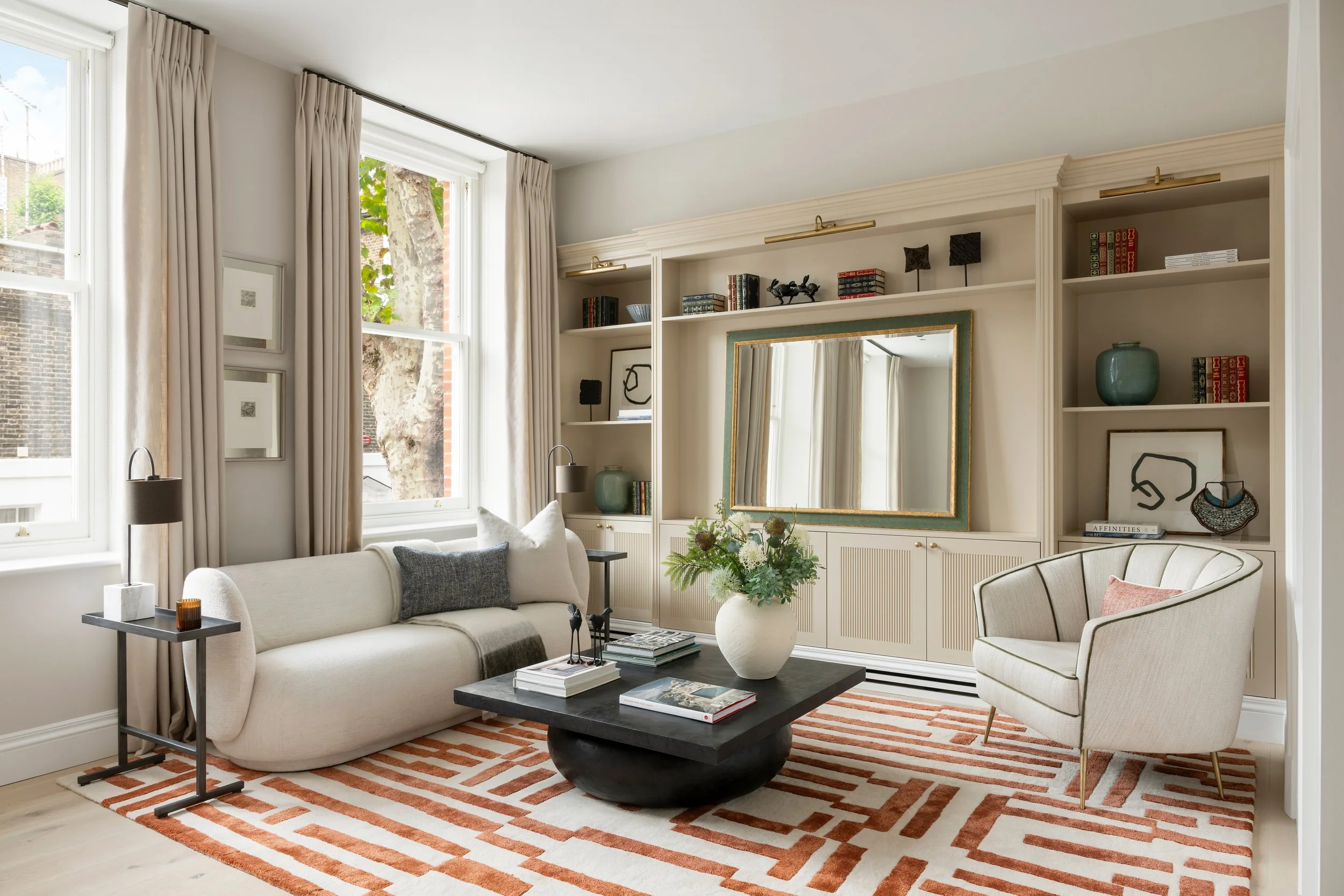 Living room with large windows, beige curtains, beige sofa with pillows, black coffee table with books and plant, white armchair, built-in bookshelves with decorative objects, mirror, and striped rug.