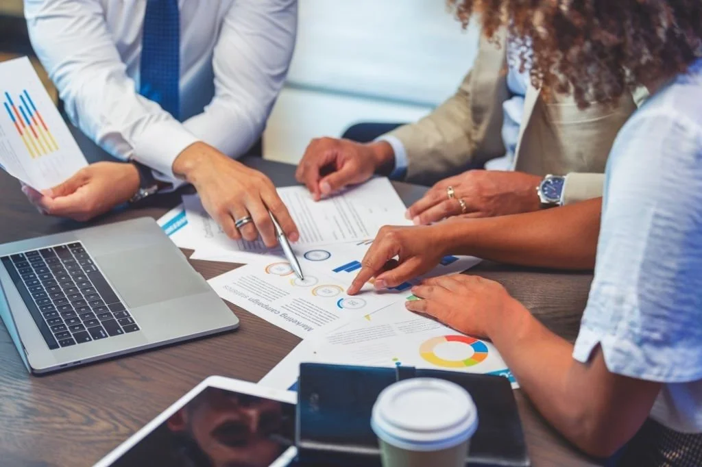 Four people discussing documents and charts at a meeting table, with a laptop, tablet, and coffee cup present.
