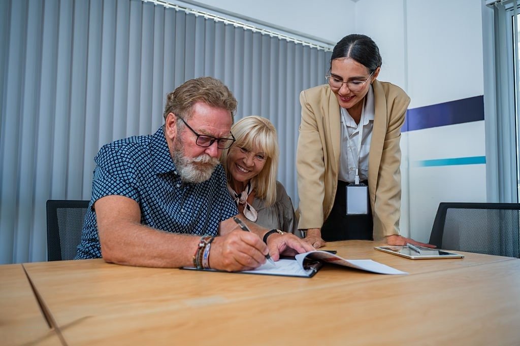 Three professionals in a meeting room, two women and a man, reviewing documents at a large wooden conference table; the man is signing papers while the women watch and smile.