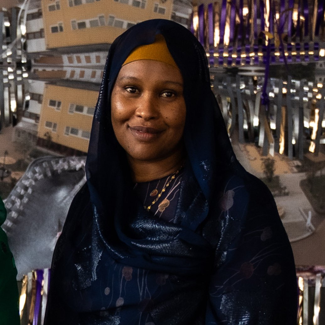Kvinna med mörk hud, bär ett mörkblått och gult huvudbonad, står på en stadsgata på kvällen med moderna byggnader i bakgrunden.
