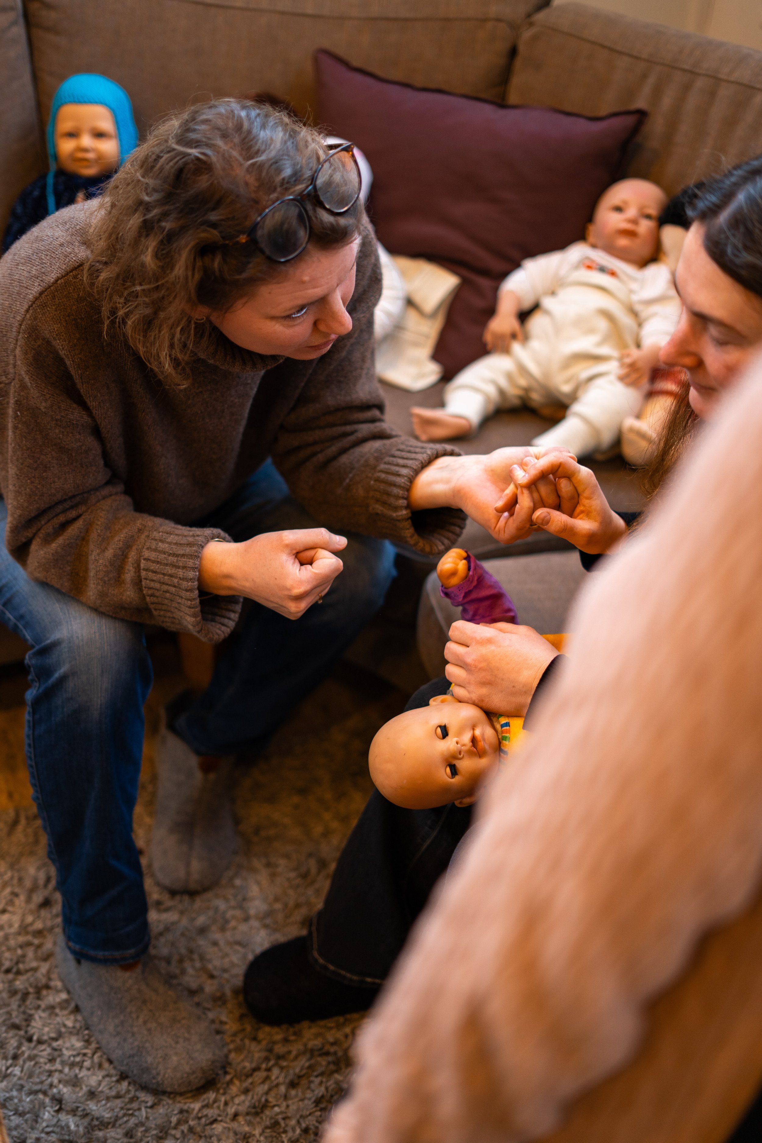 En vuxen kvinna böjer sig fram för att prata med en babydocka, medan en annan kvinnas hand håller dockan. I bakgrunden ligger två babydockor på en soffa, en med blå hatt och en med vit klädsel. Personen är inomhus på ett vardagsrumgolv.