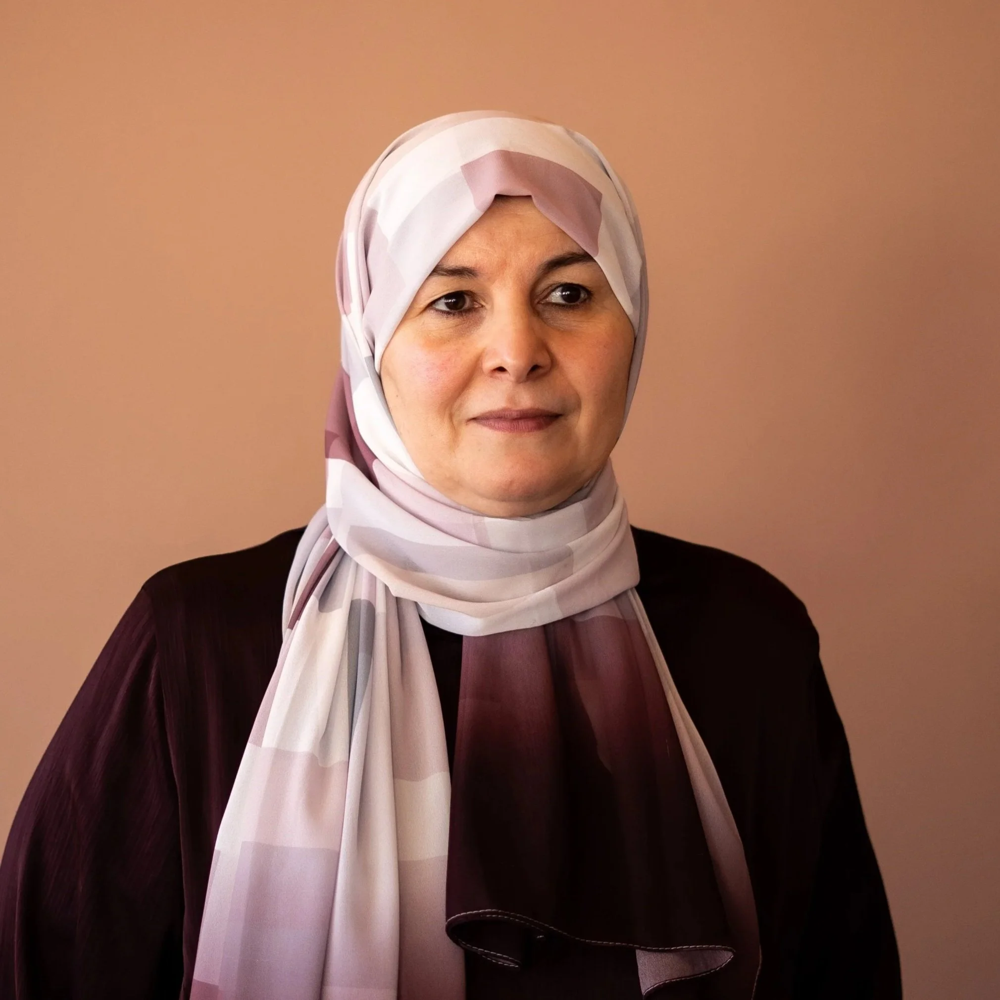 Närbild av en kvinna med ljus hy och mörka ögon, bär en rosa och lila mönstrad hijab och en svart tröja, står mot en solid ljusbrun bakgrund.