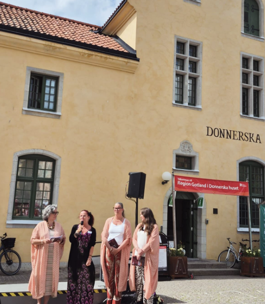 Fyra kvinnor står utanför en gul byggnad, med en röd banderoll som hälsar välkommen till regionen Gotland och Donnerska huset. En av kvinnorna talar i mikrofon, andra lyssnar. Två cyklar är parkerade i närheten.