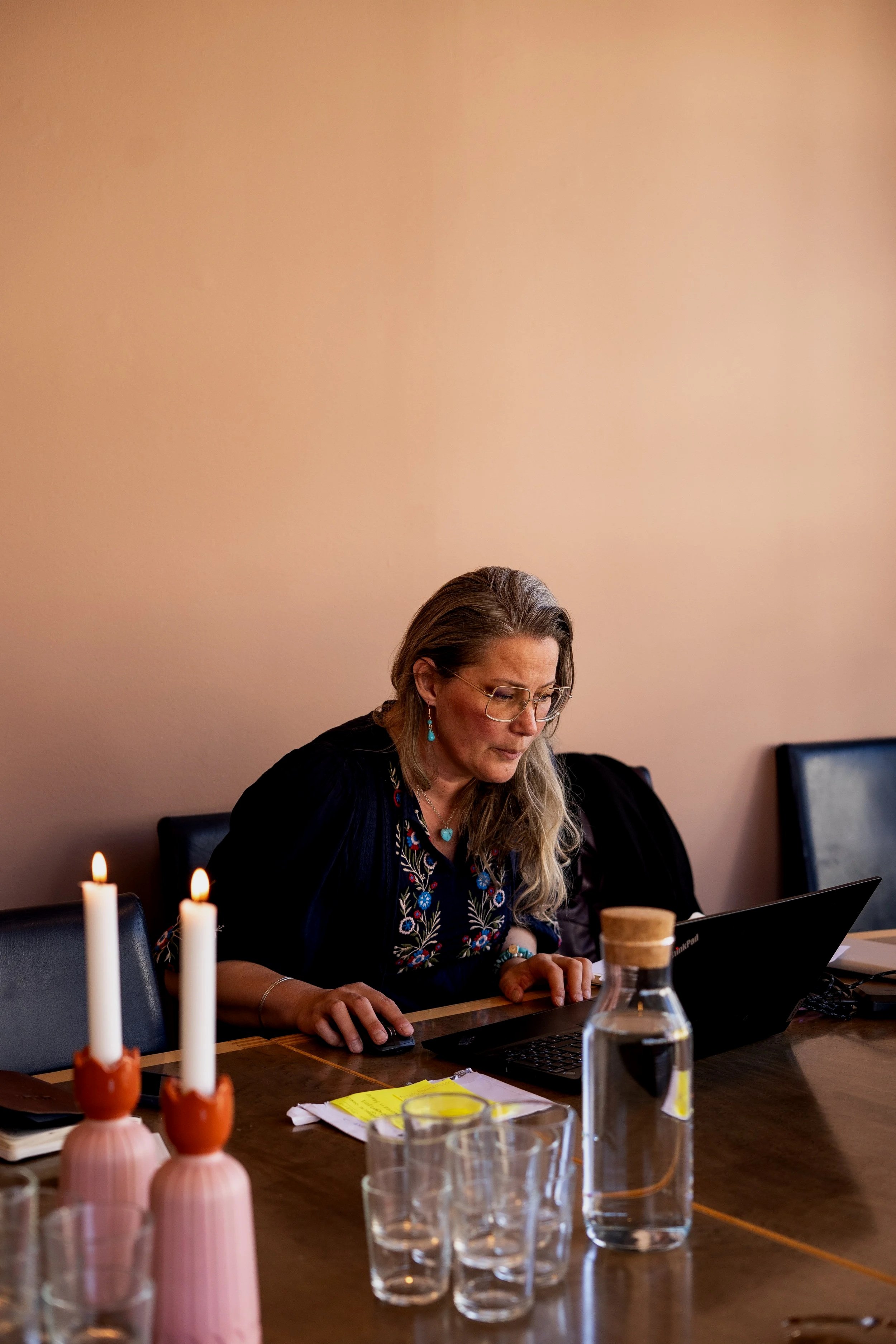 En kvinna med lång, blond och grå hår, bär glasögon och ett svart plagg samt smycken, sitter vid ett bord med en laptop. På bordet finns även ett vatten samt ett avslappnat ljus i förgrunden.