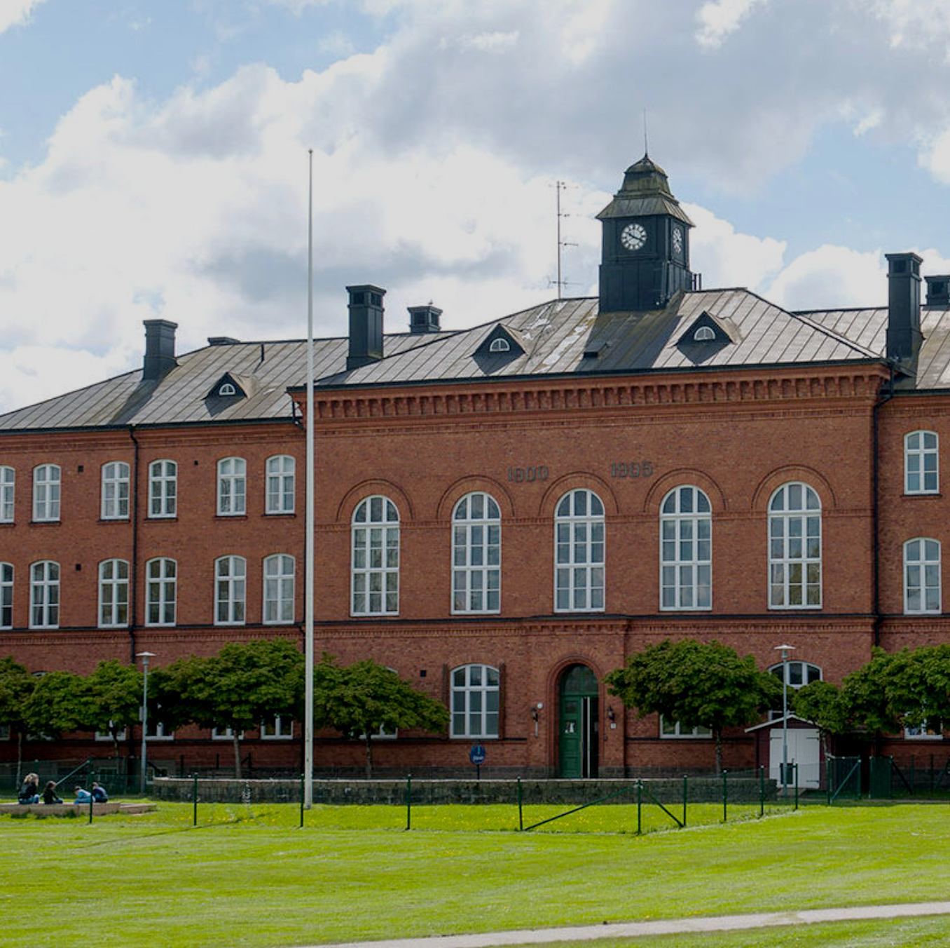 En röd tegelbyggnad med stora fönster och ett klocktorn, omgiven av grön gräsmatta och några träd, under en molnig himmel.