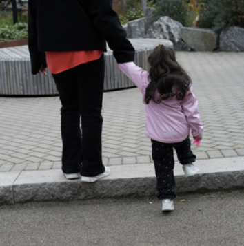 En vuxen håller handen på en liten flicka som går på en trottoar i en park eller offentlig plats.